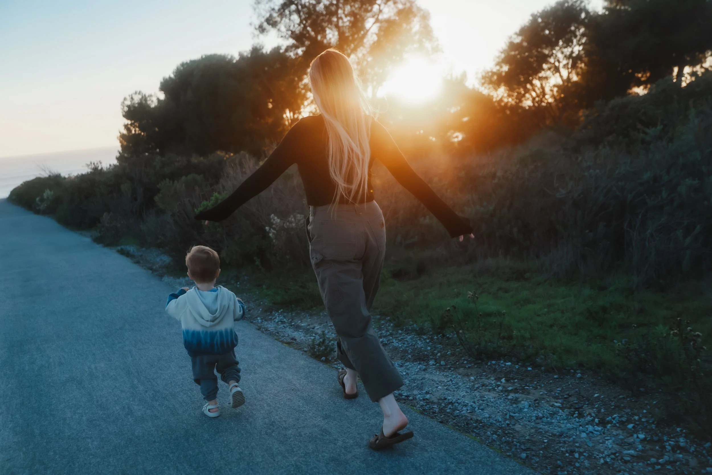 A woman and a young boy walking on a paved path outdoors during sunset, with the woman extending her arms and the boy running ahead.