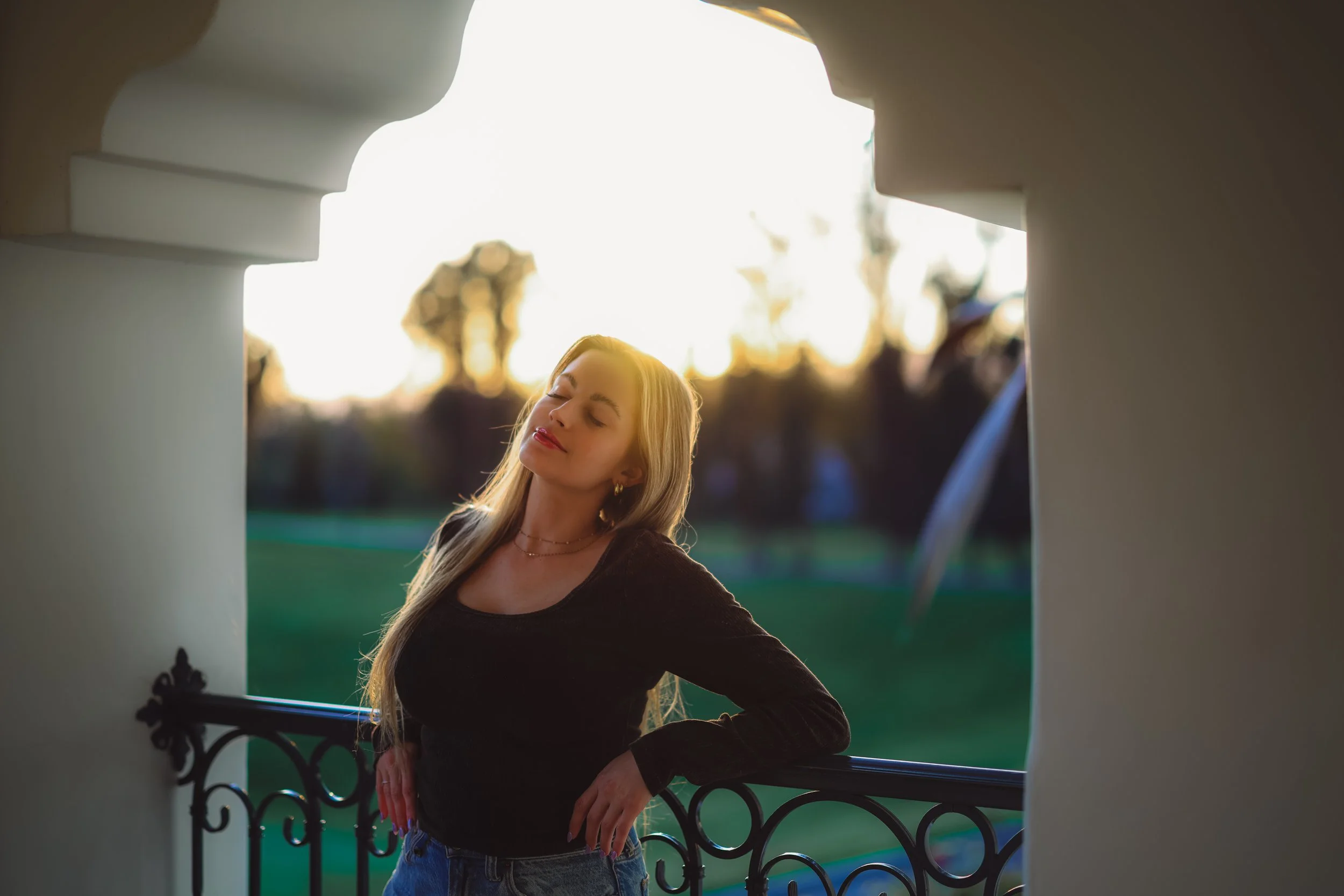 Woman with long hair in a black top and jeans standing on a balcony with eyes closed during sunset.