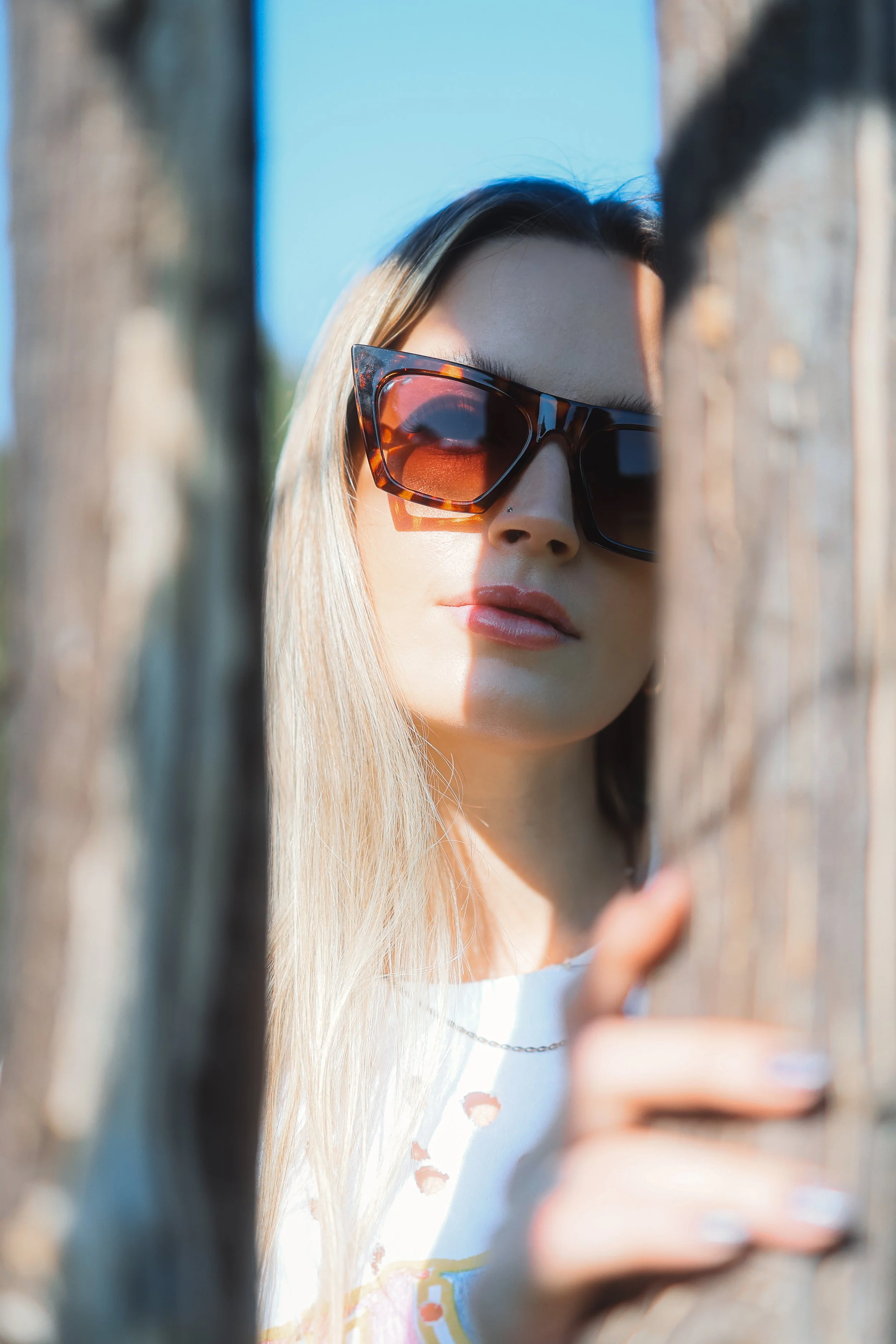 A woman wearing large, tortoiseshell sunglasses and a white t-shirt with a colorful print, looking through a gap in a wooden fence, with blond hair and a neutral expression.