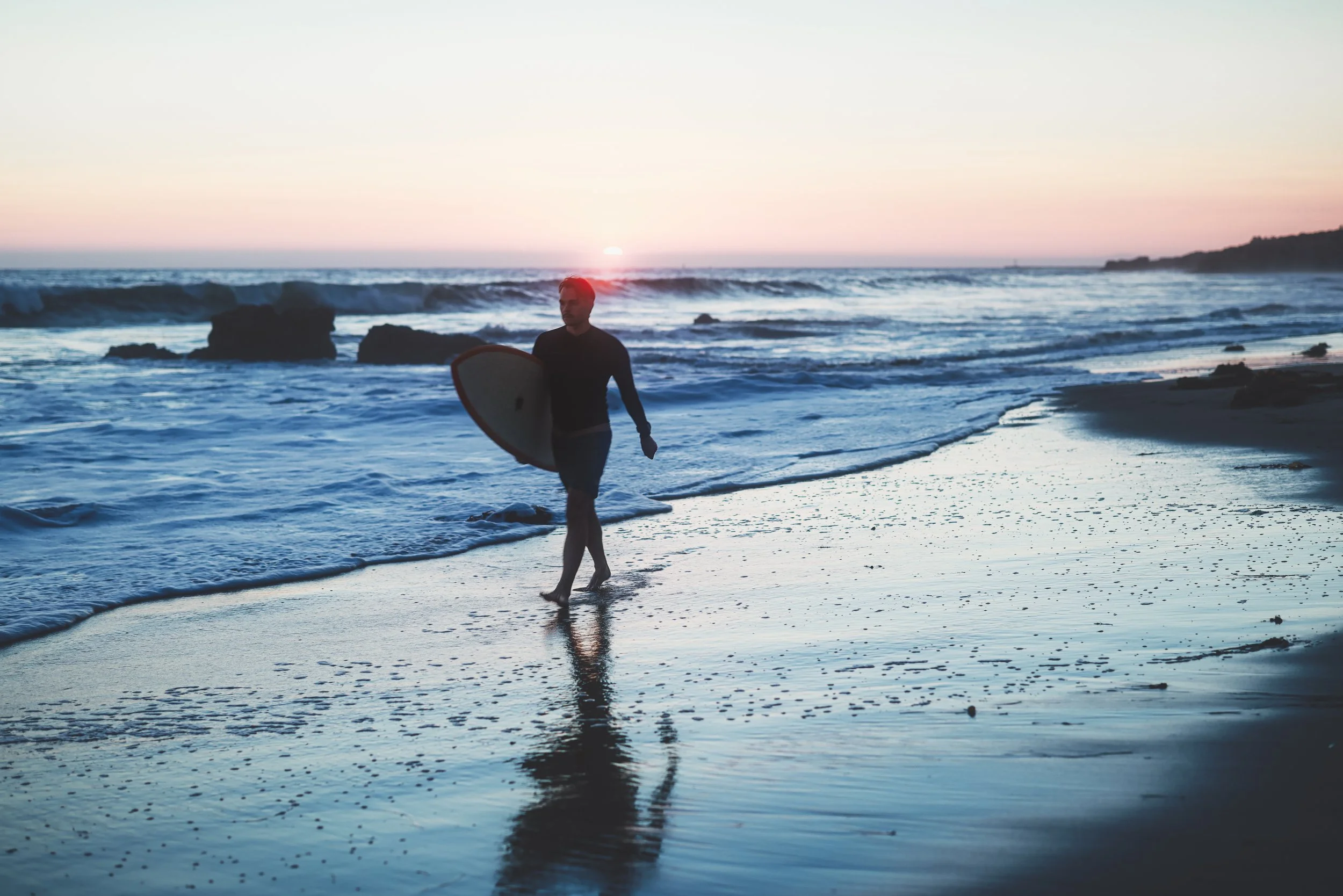 A person walks barefoot along the sandy shoreline holding a surfboard with the sunset in the background.