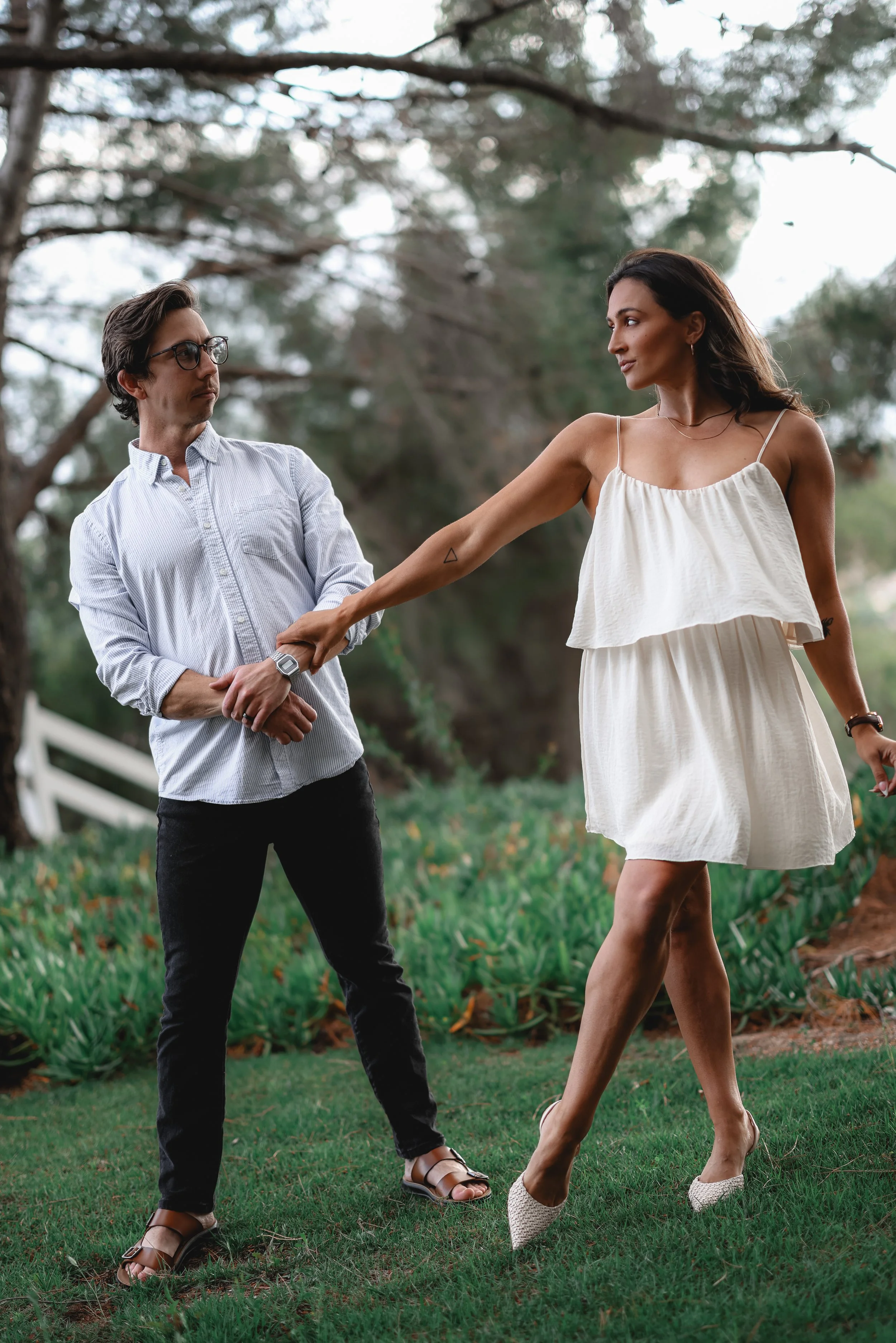 A man in glasses and a woman in a white dress holding hands in a park with trees and grass.
