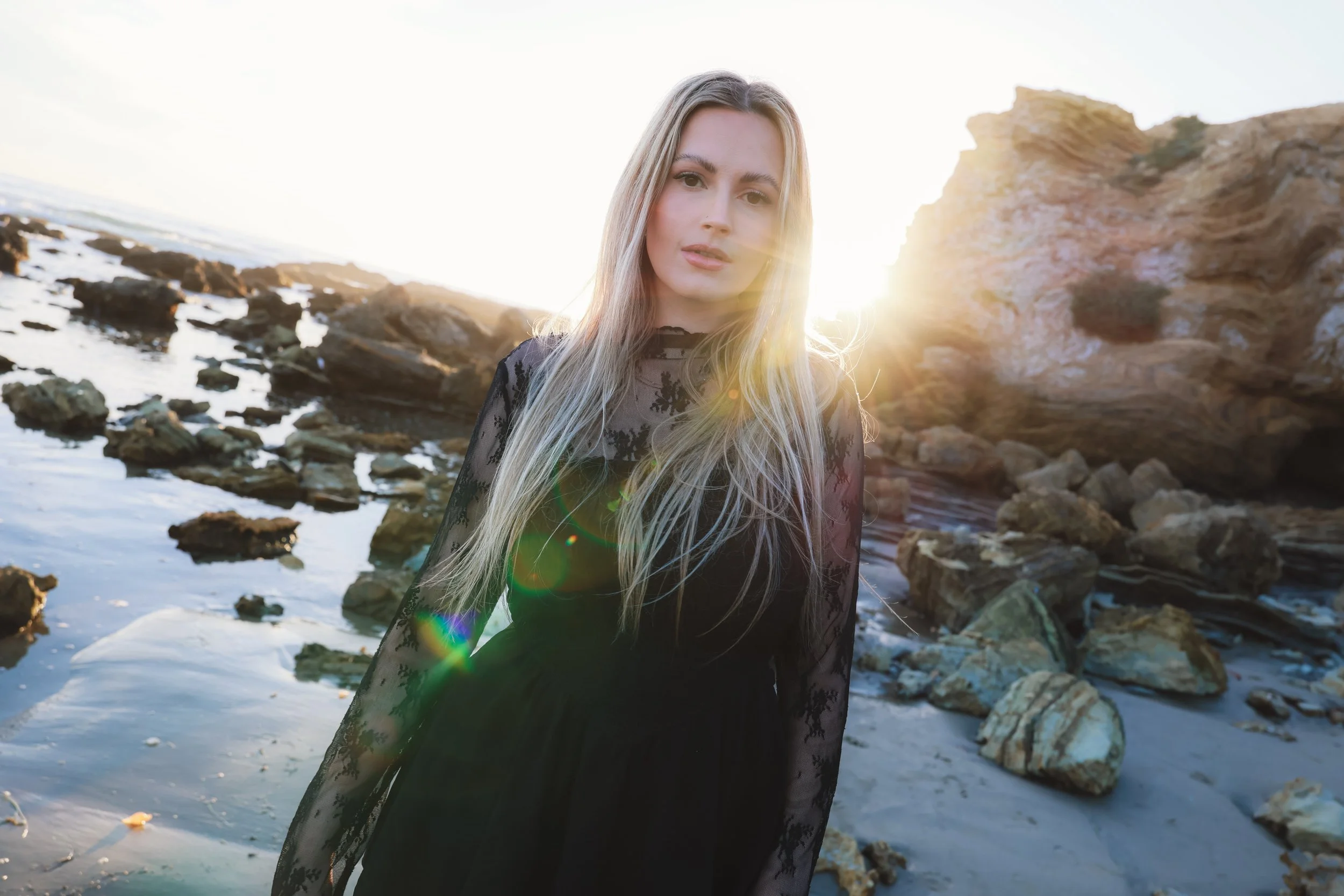 Young woman in a black lace dress standing on a rocky beach at sunset with sunlight shining behind her.