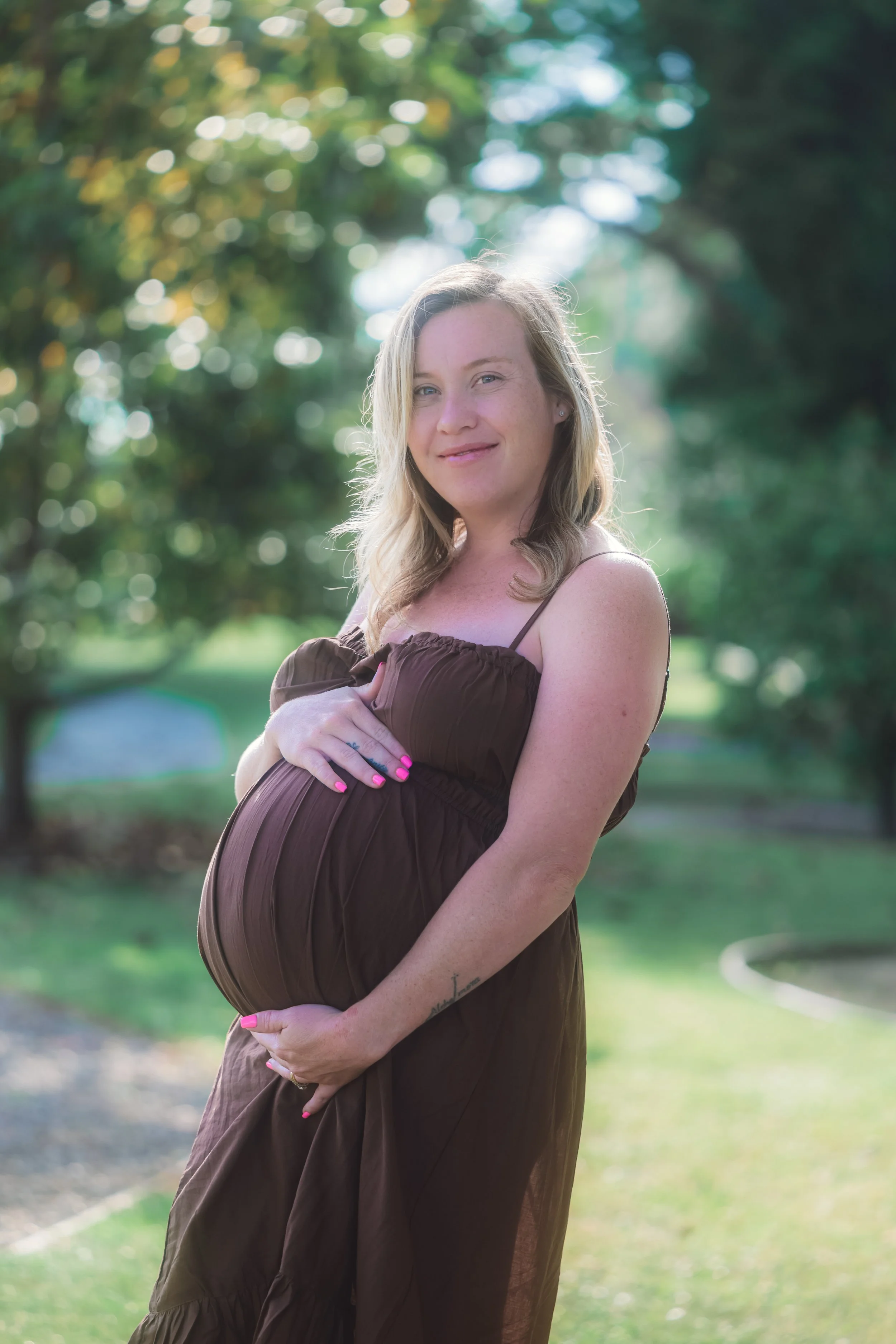 Pregnant woman in a brown dress standing outdoors in a park, holding her belly and smiling gently, with blurred trees and greenery in the background.