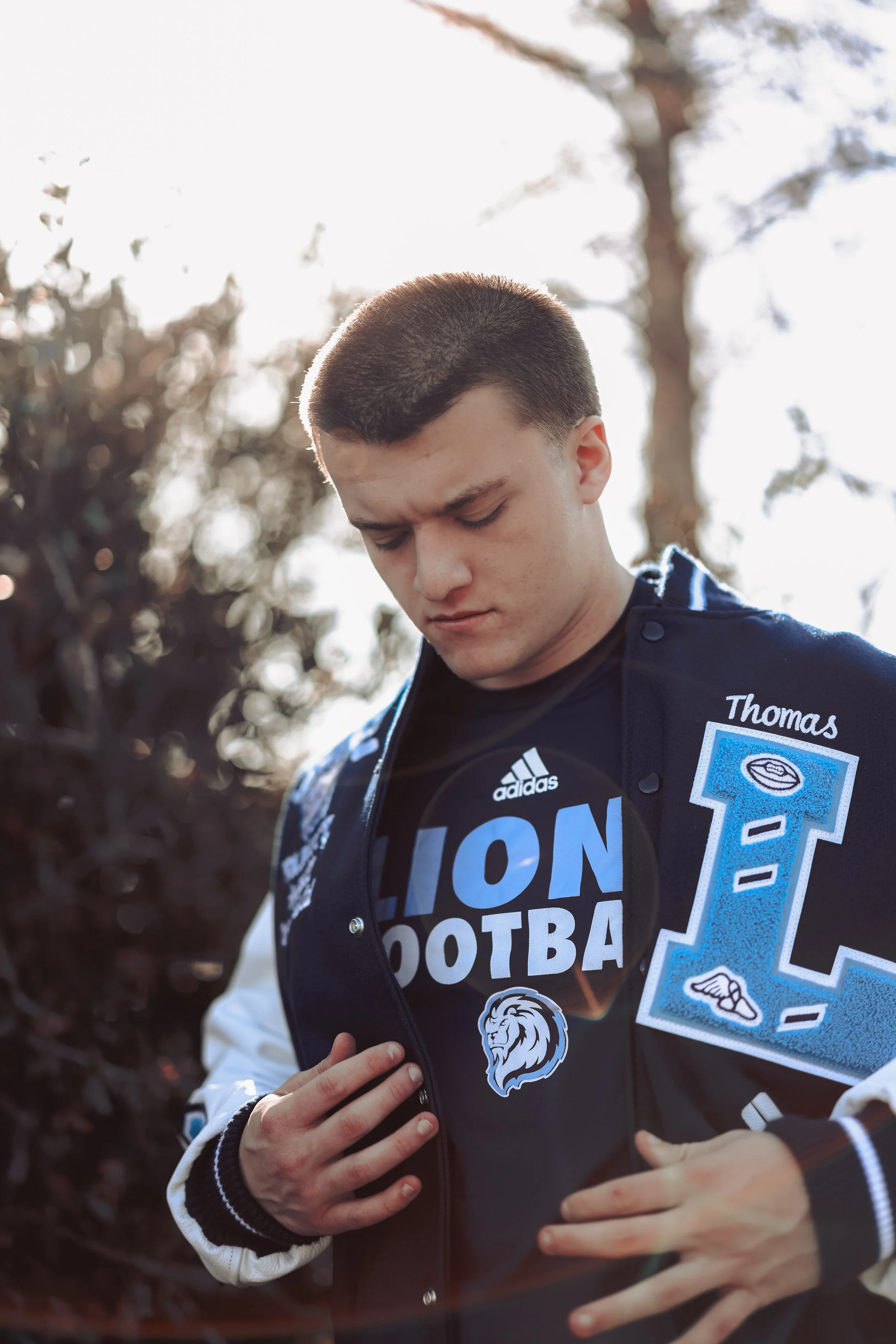 Young man looking down, wearing a black athletic shirt with blue and white lettering and a varsity-style jacket with patches, outdoors on a sunny day.