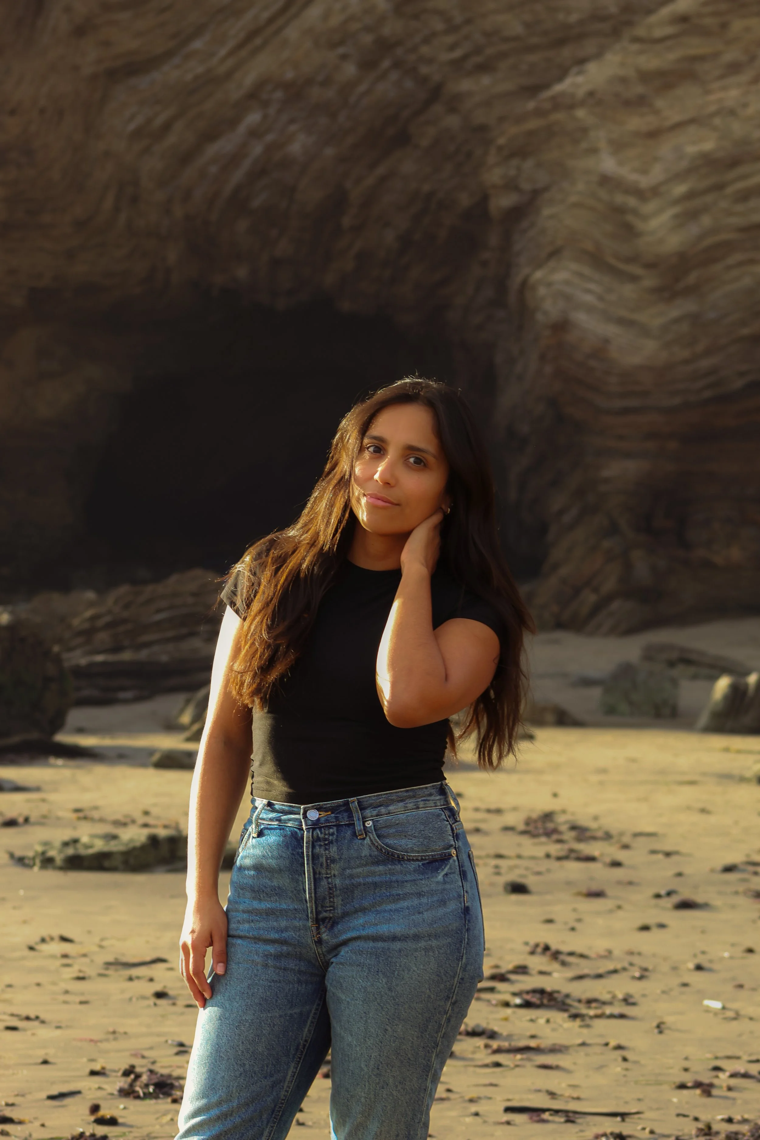 A woman standing on the beach in front of a large rock formation with a cave, during sunset.