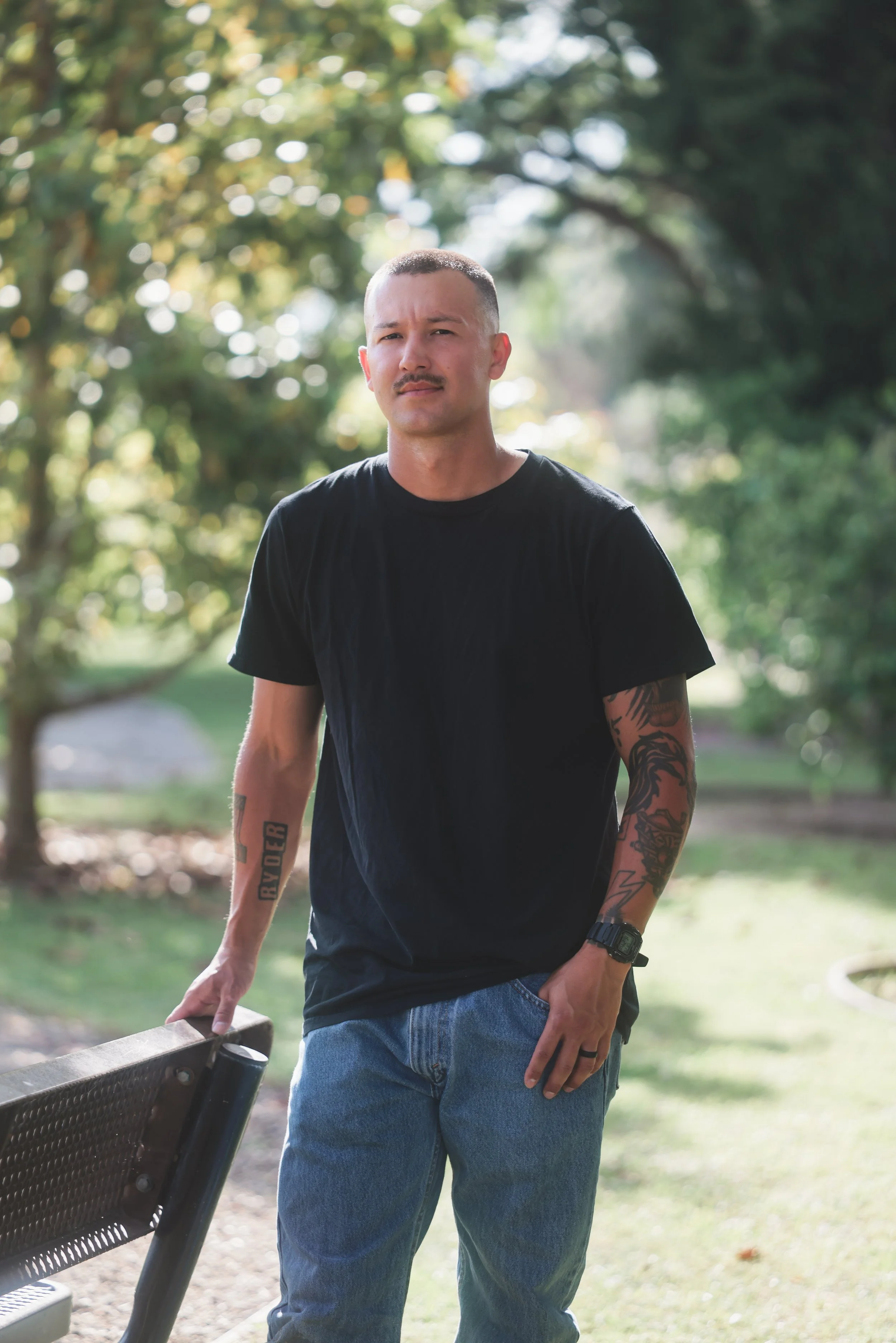 A young woman with long blonde hair stands outdoors near a railing and a large bush, dressed in a black long-sleeve shirt and blue jeans, with the setting sun behind her.