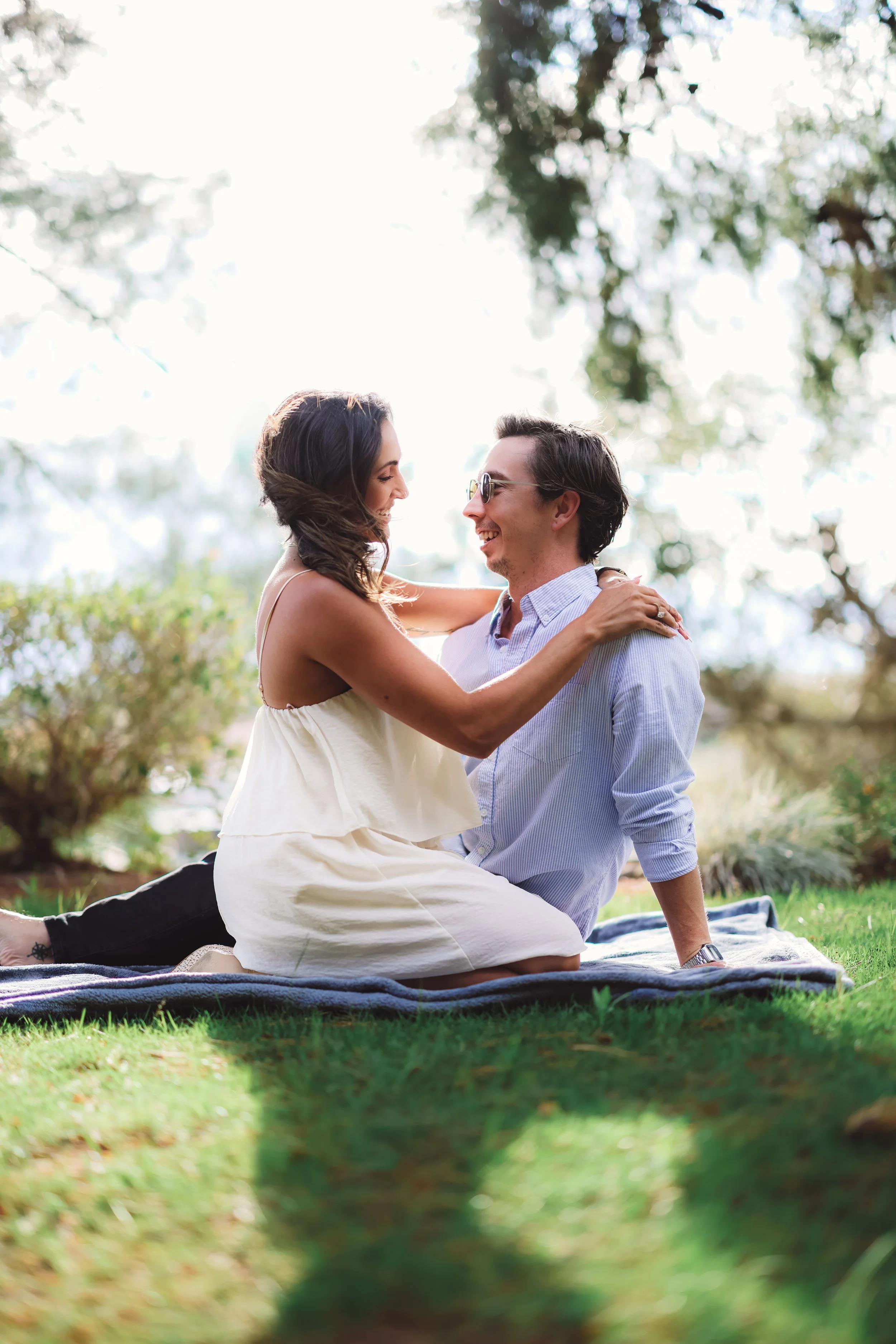 A couple sitting on a blanket outdoors, facing each other and smiling, surrounded by trees and greenery, with sunlight filtering through the leaves.