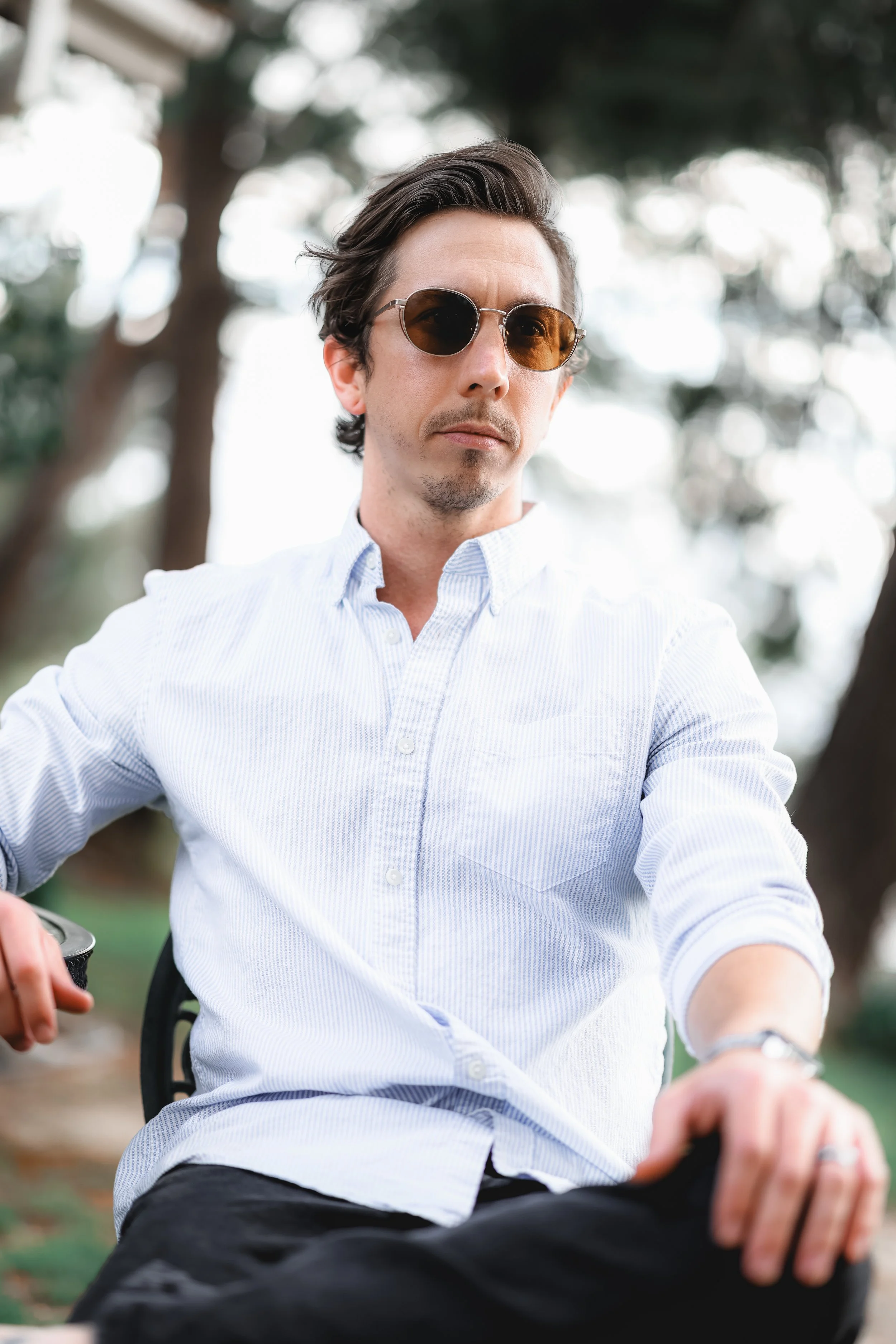 A man with dark hair wearing sunglasses and a light blue button-up shirt sitting outdoors, with trees and bright sky in the background.