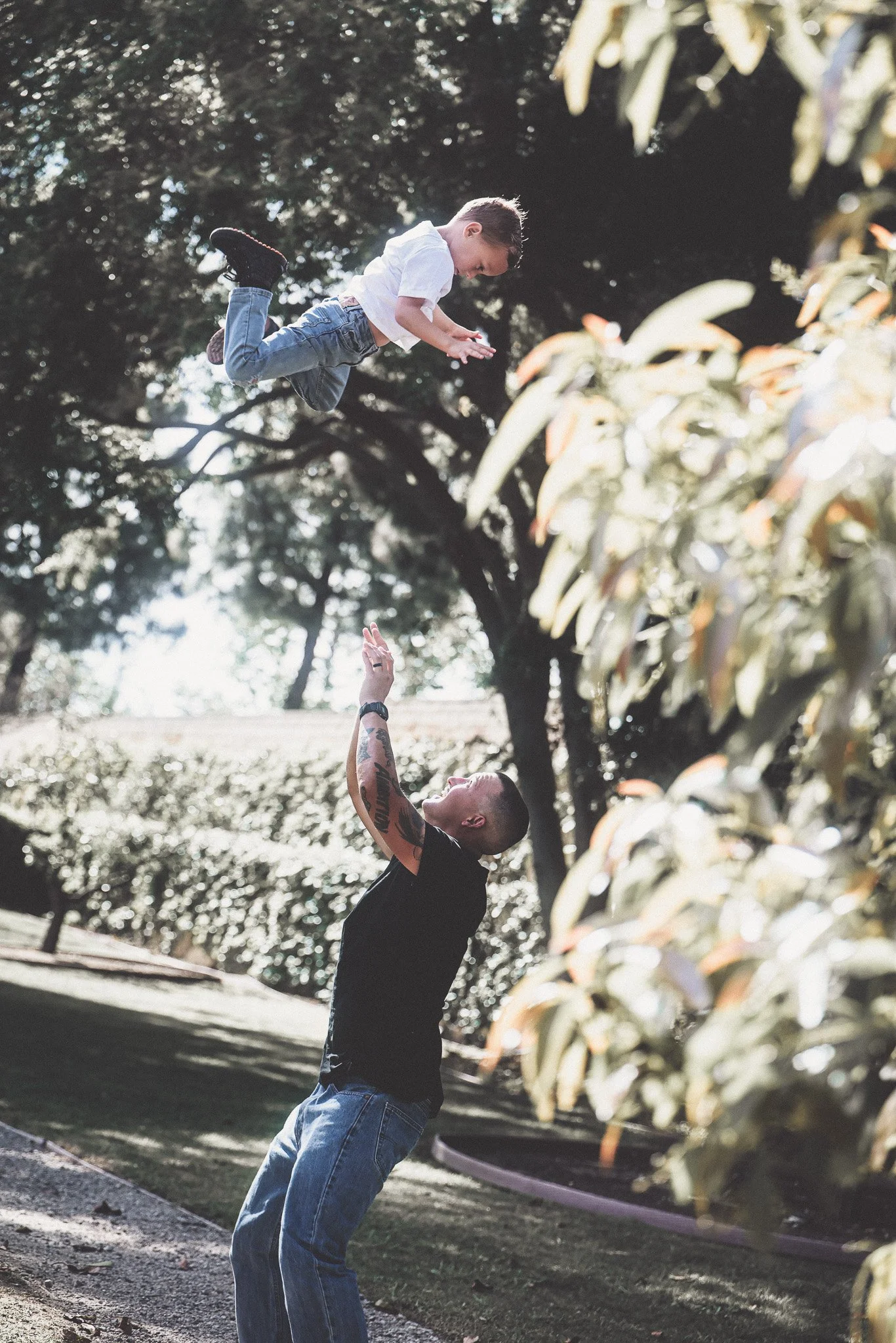 A man is playing with a young boy outdoors, tossing him into the air. The man is smiling and looking up at the boy, who is mid-air with arms extended downward, surrounded by trees.