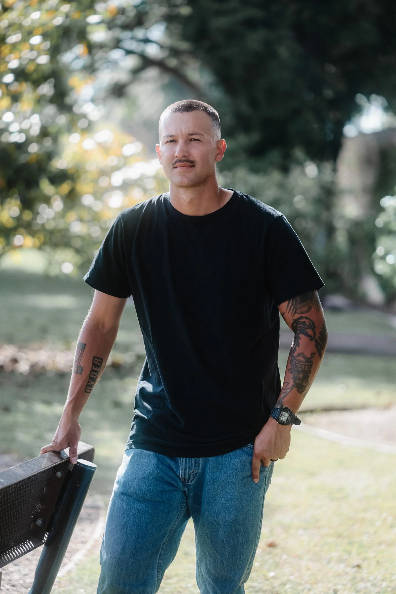 A young man with a shaved head and tattoos on his arms, wearing a black T-shirt and jeans, holding a lawn mower handle in an outdoor park setting with trees.