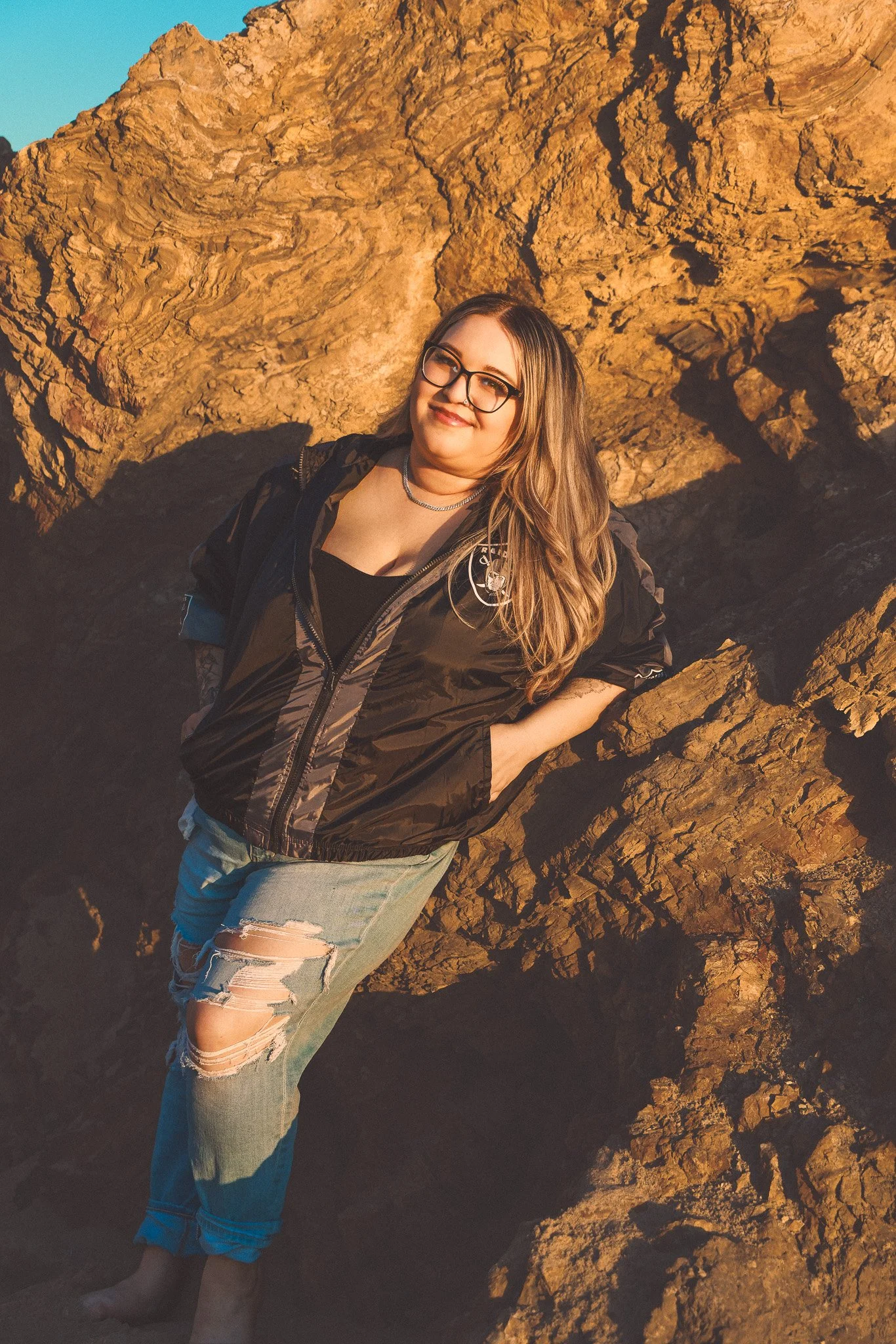 A woman with glasses and long hair standing next to a large rock formation during sunset, smiling and dressed in a black jacket and ripped jeans.