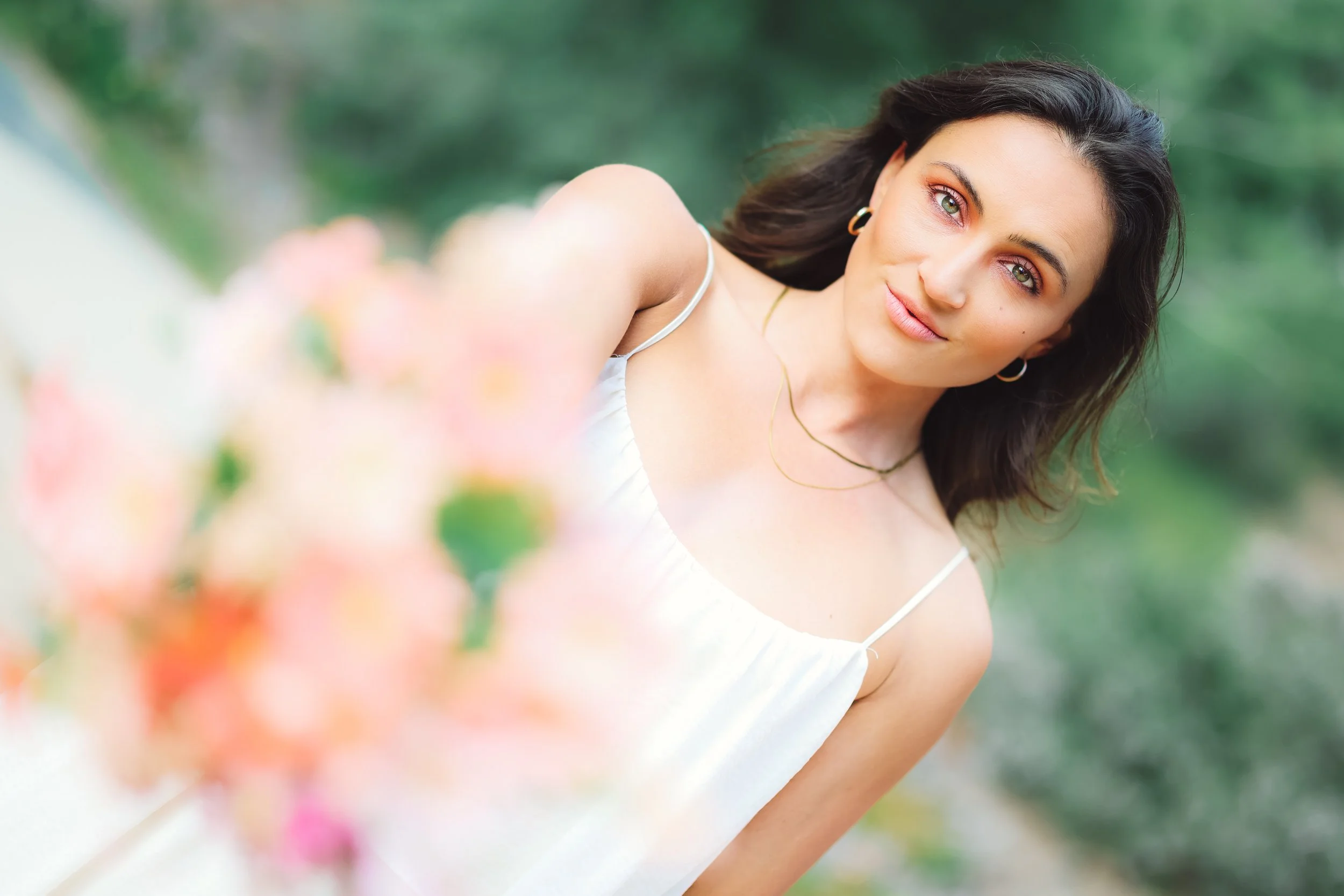 A woman with dark hair and green eyes wearing a white sleeveless dress, standing outdoors with blurred flowers in the foreground and greenery in the background.