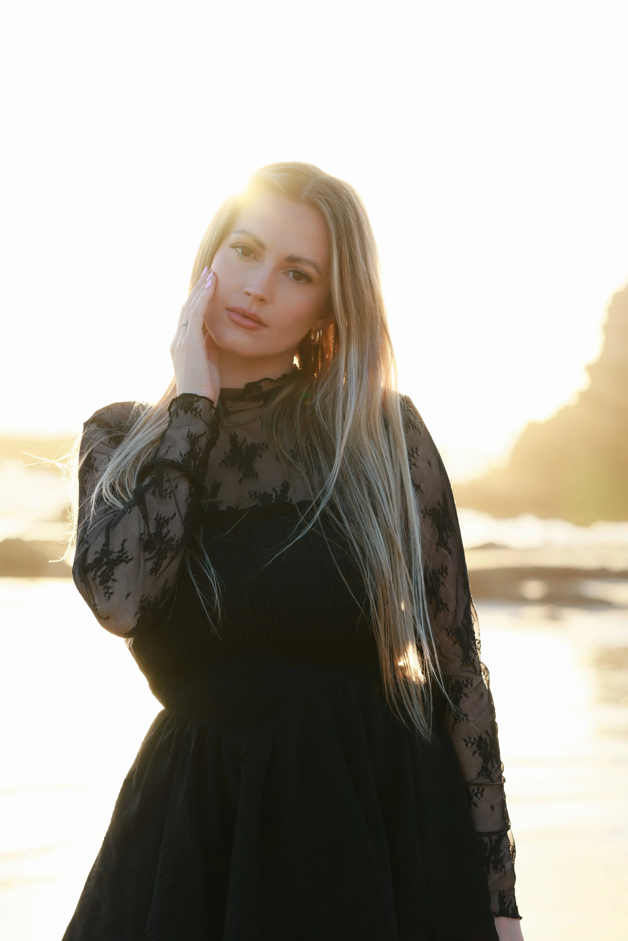 A young woman with long blonde hair wearing a black lace dress, standing outdoors at sunset near the beach.