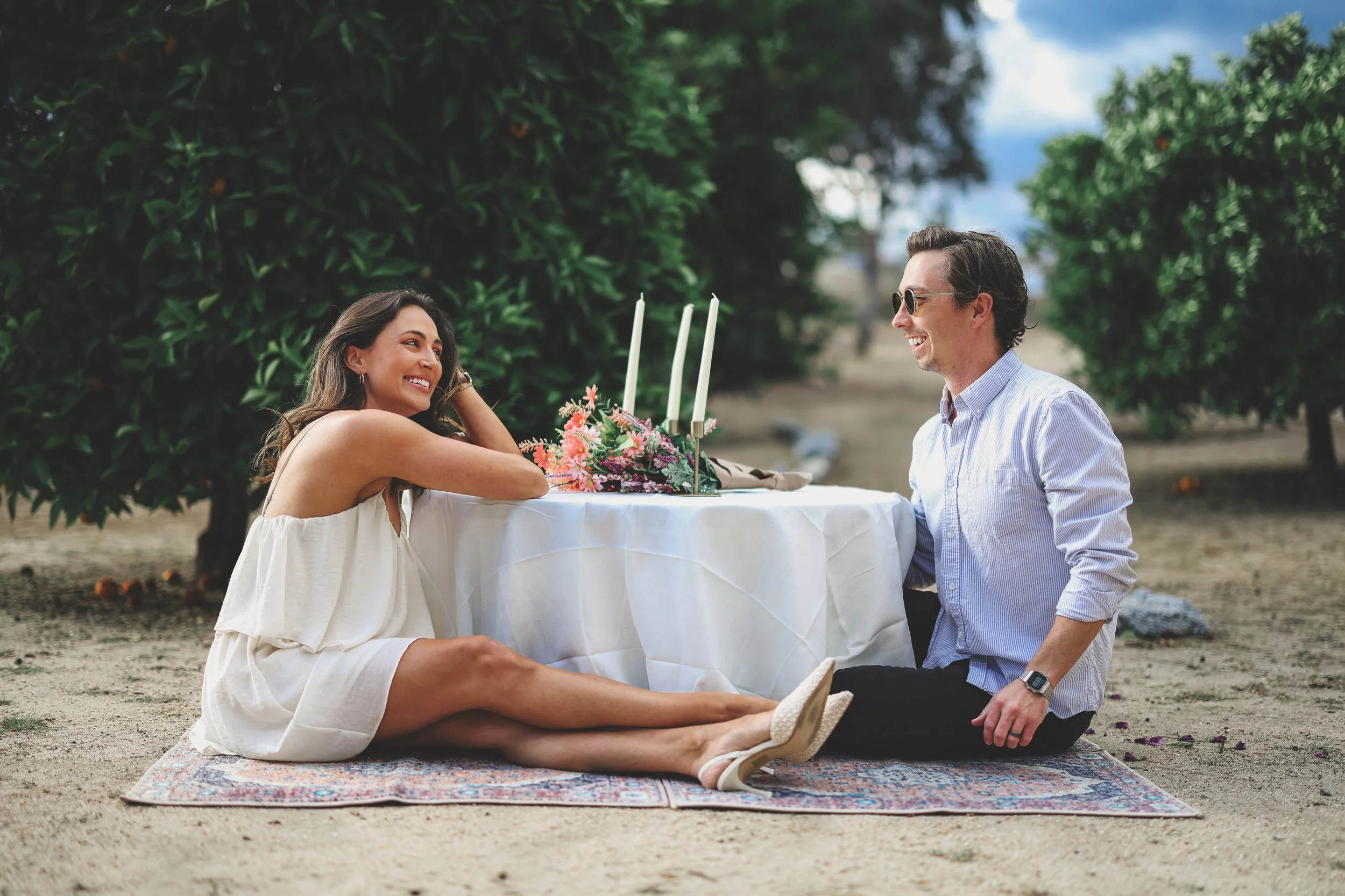 A couple sitting on a colorful rug outdoors, with a table set with pink flowers and candles, smiling and enjoying a romantic moment on a partly cloudy day with trees in the background.