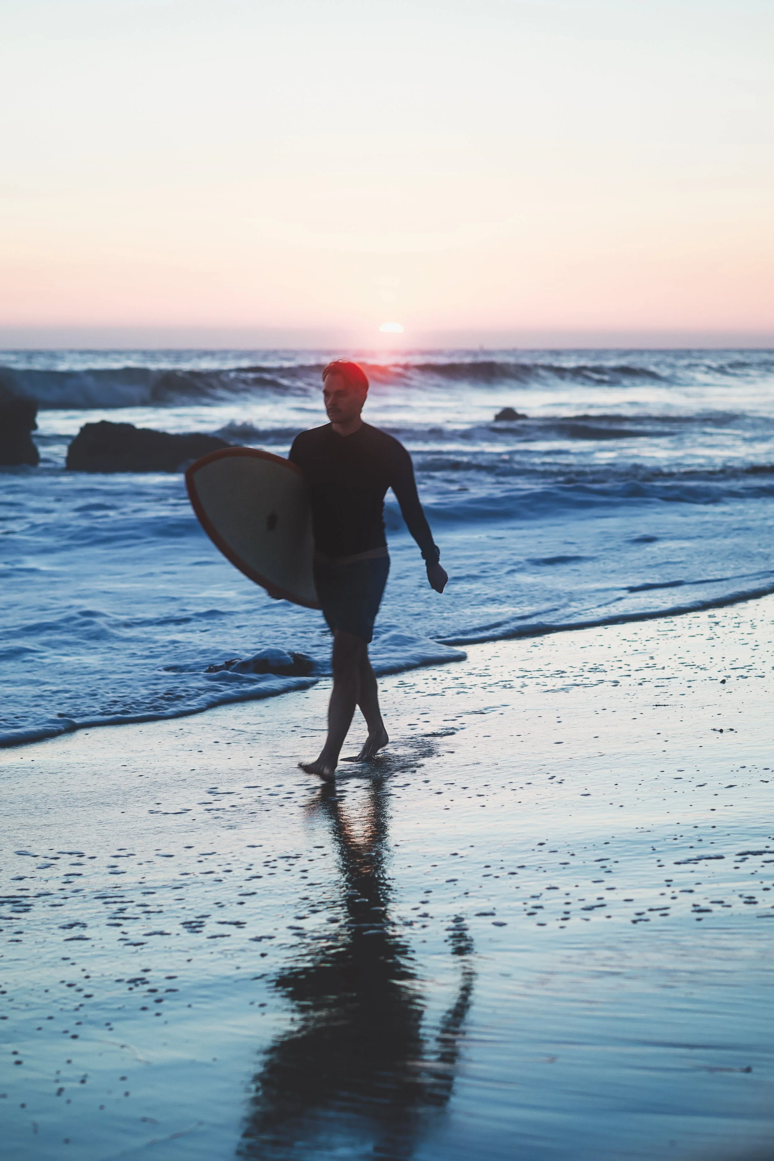 A man carrying a surfboard walking along the beach at sunset with waves in the background.
