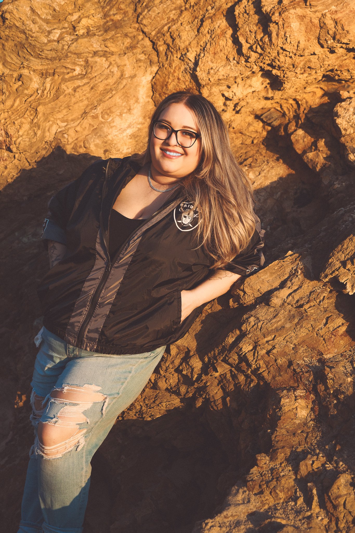 A woman with glasses, long light brown hair, and a black jacket with a patch on the sleeve, standing outdoors in front of a large rock formation, smiling at the camera.