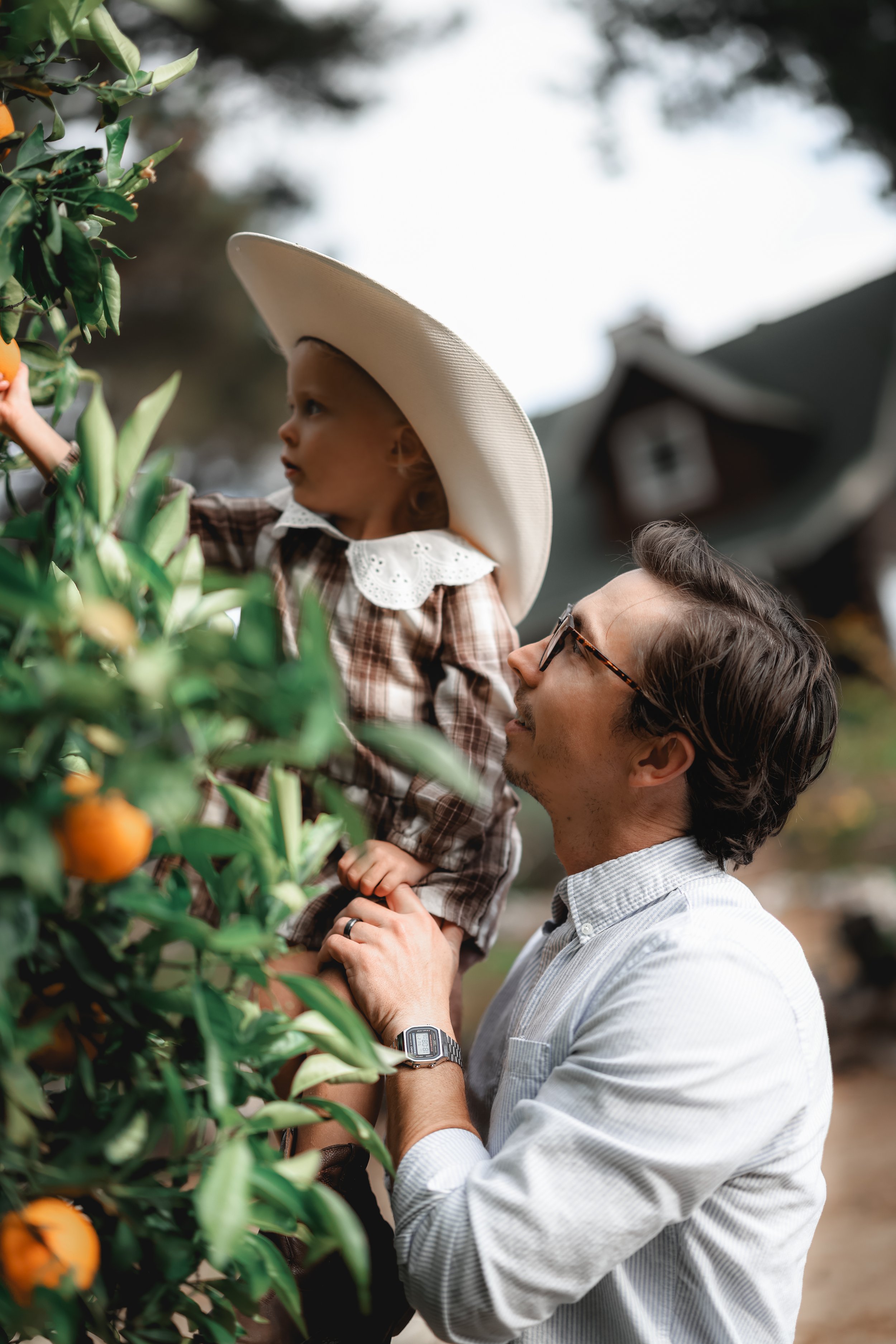 A man with glasses and a child wearing a large sunhat and plaid shirt picking oranges from a tree.