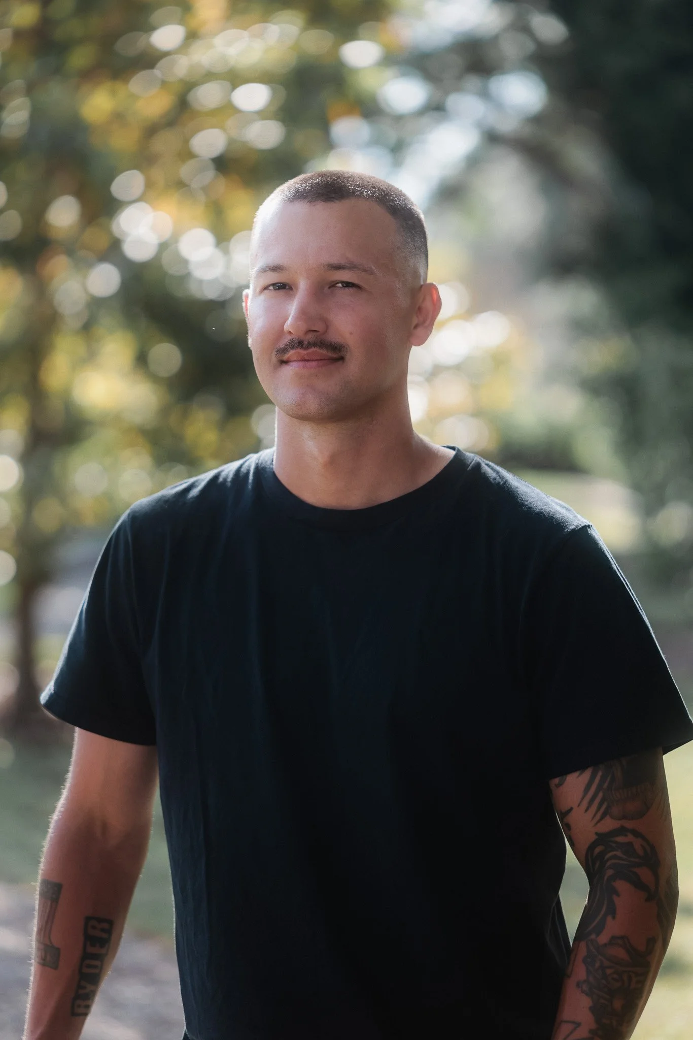 A woman with long blonde hair, wearing a black top and gold jewelry, standing outdoors during sunset with a blurred background of trees and sky.