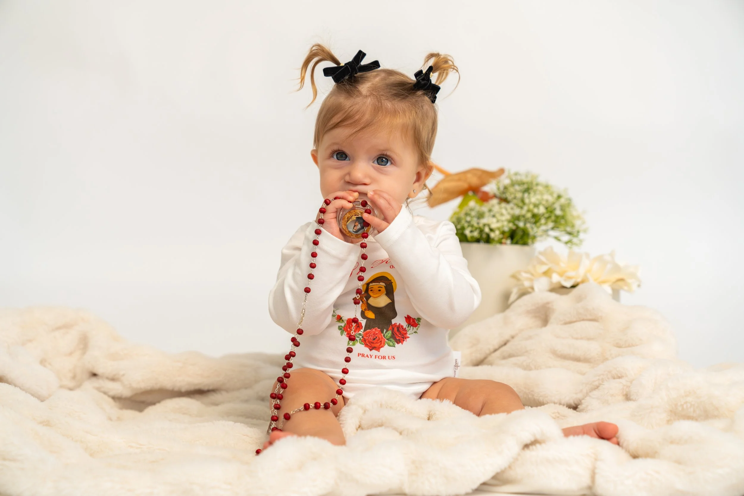 Young girl with pigtails sitting on a plush blanket, holding a rosary and a religious medal, with a vase of flowers in the background.