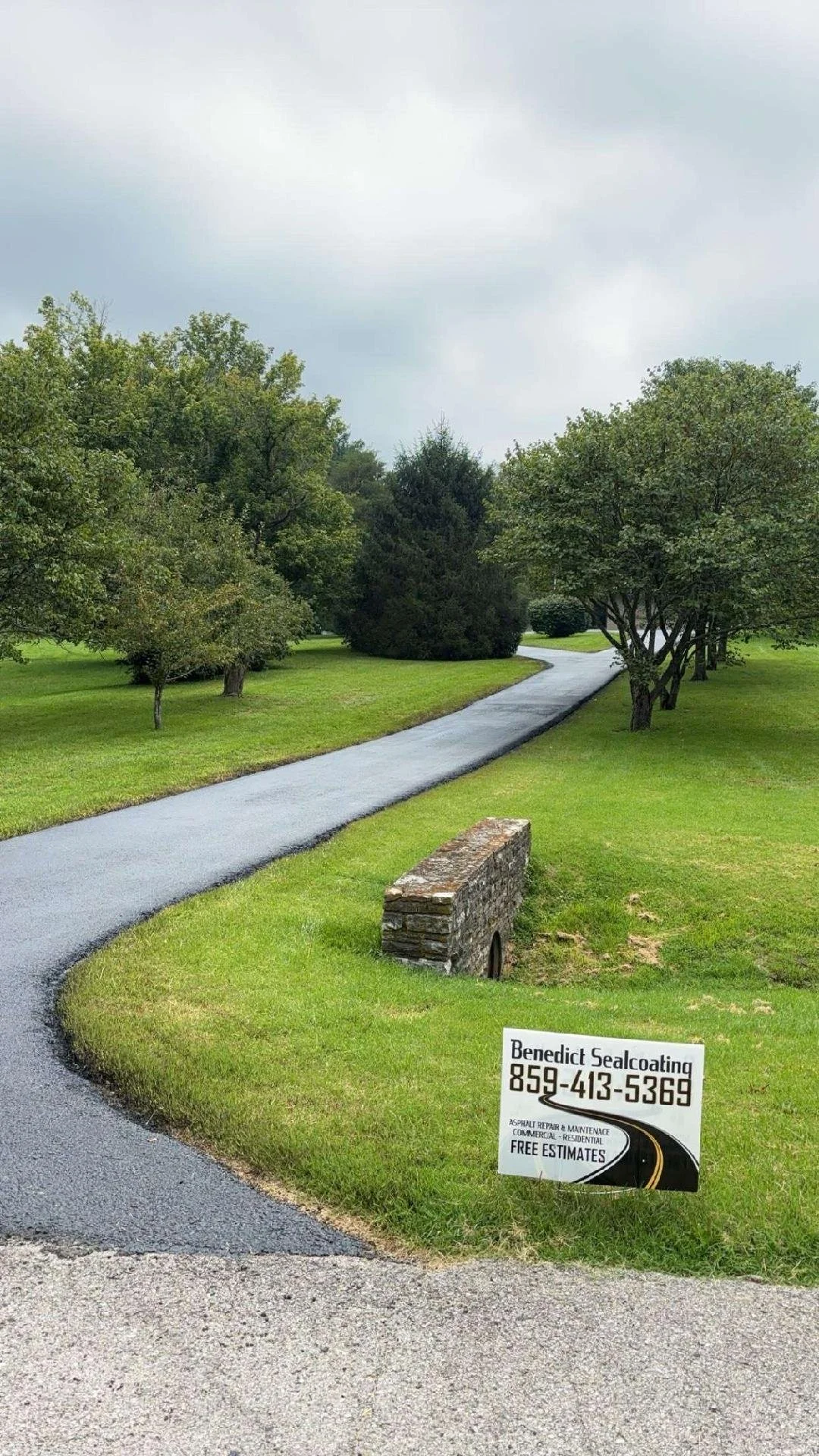 A asphalt driveway surrounded by green grass and trees, with a sign advertising Benedict Sealcoating and a small brick structure at the edge.