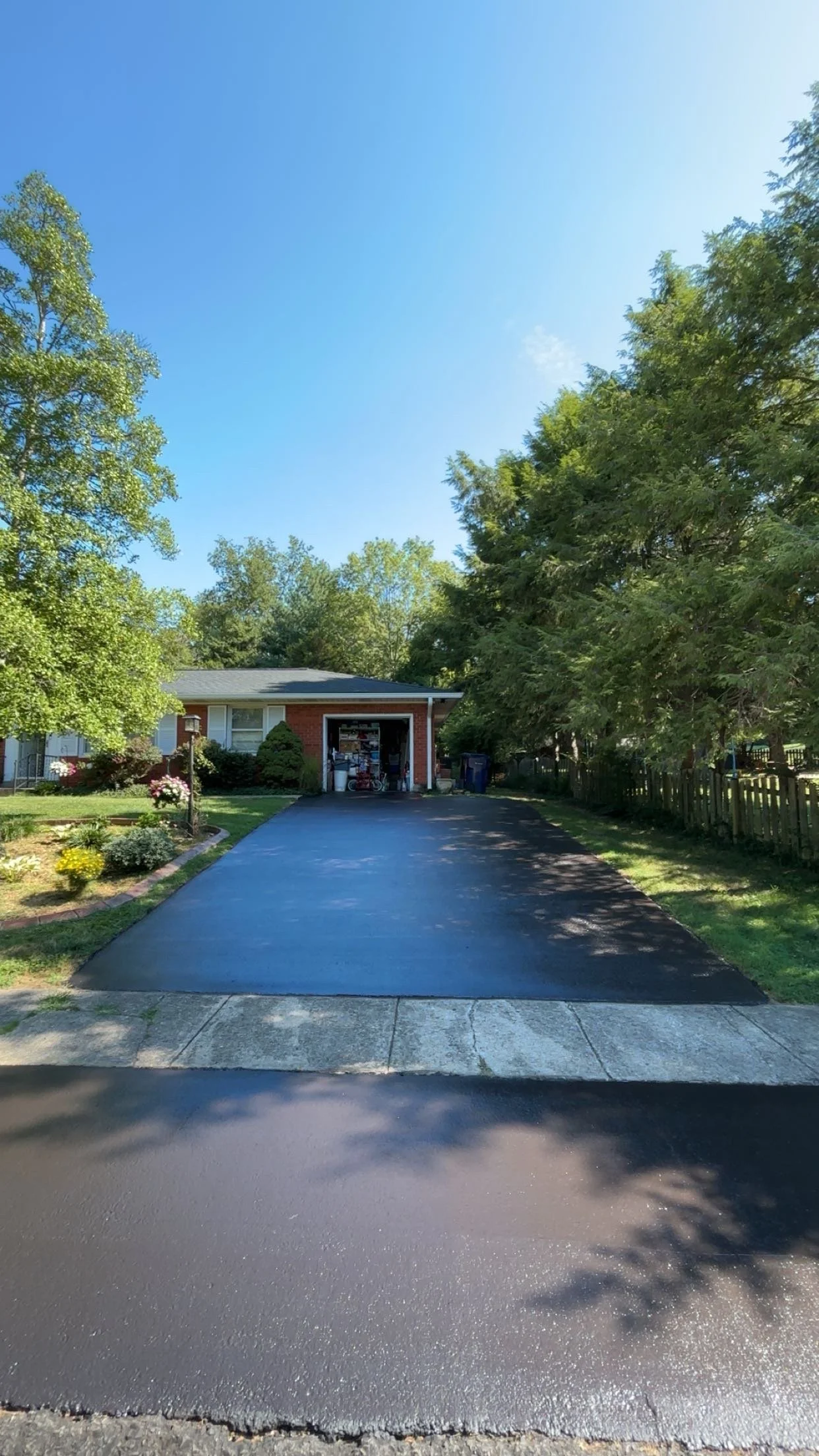 A driveway leading to a brick garage attached to a house, with a well-maintained yard and trees on either side, under a clear blue sky.