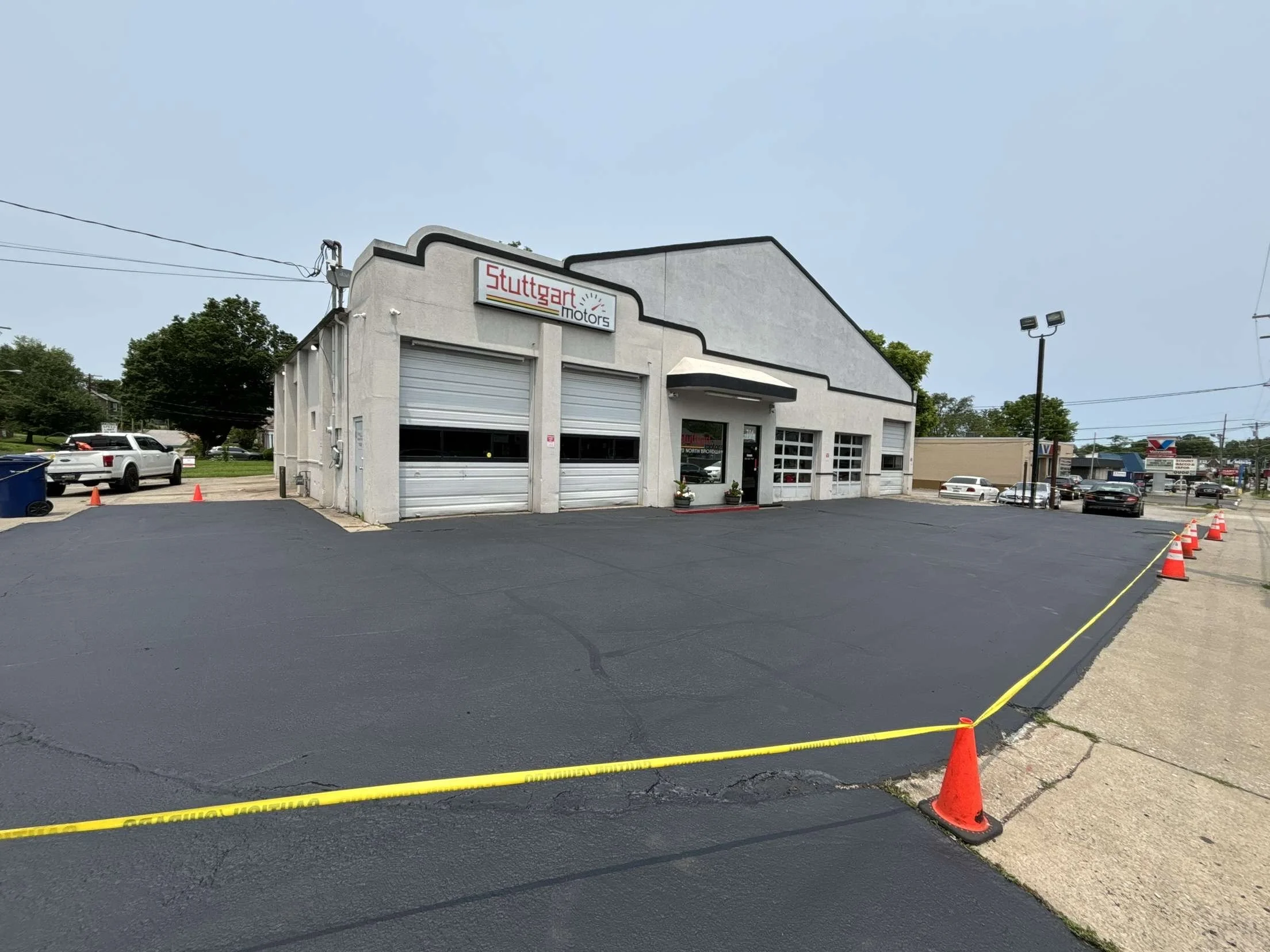 A car dealership building with a sign that reads 'Stuttgart Motors', several garage doors, and a small entrance door, with a freshly sealcoated paved parking lot cordoned off with orange traffic cones and yellow caution tape
