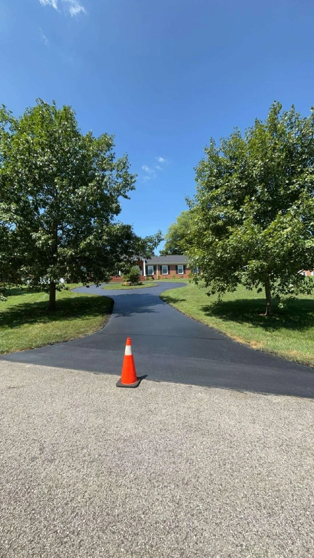 A blacktop driveway with a traffic cone blocking entry, leading to a house with a brick exterior and a front porch, flanked by green trees under a clear blue sky.