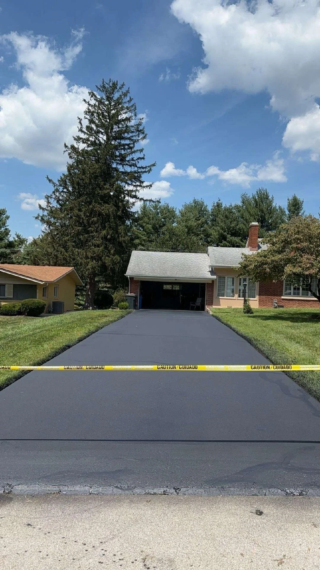 Freshly Sealcoat paved driveway with caution tape across it in front of a house, with grass on either side, trees, and a partly cloudy sky.