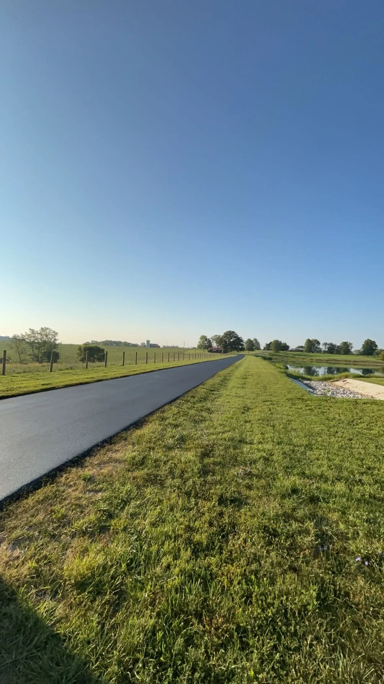 A sealcoated country farm road running through a grassy countryside with a fence on one side and a small pond on the other, under a clear blue sky.