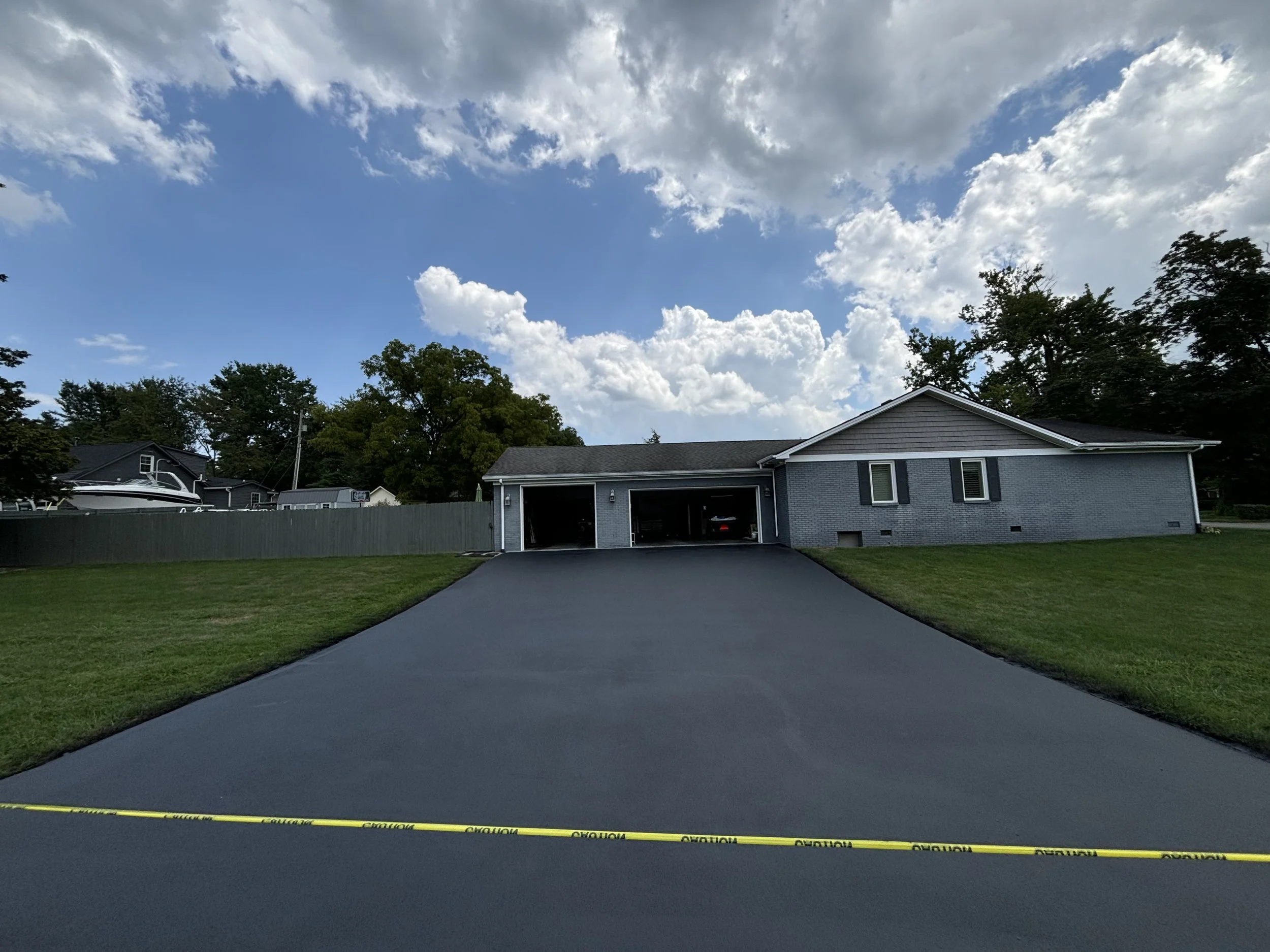 A newly paved driveway leading to a gray house with an attached garage, green grass on either side, a metal fence, and a partially cloudy blue sky.