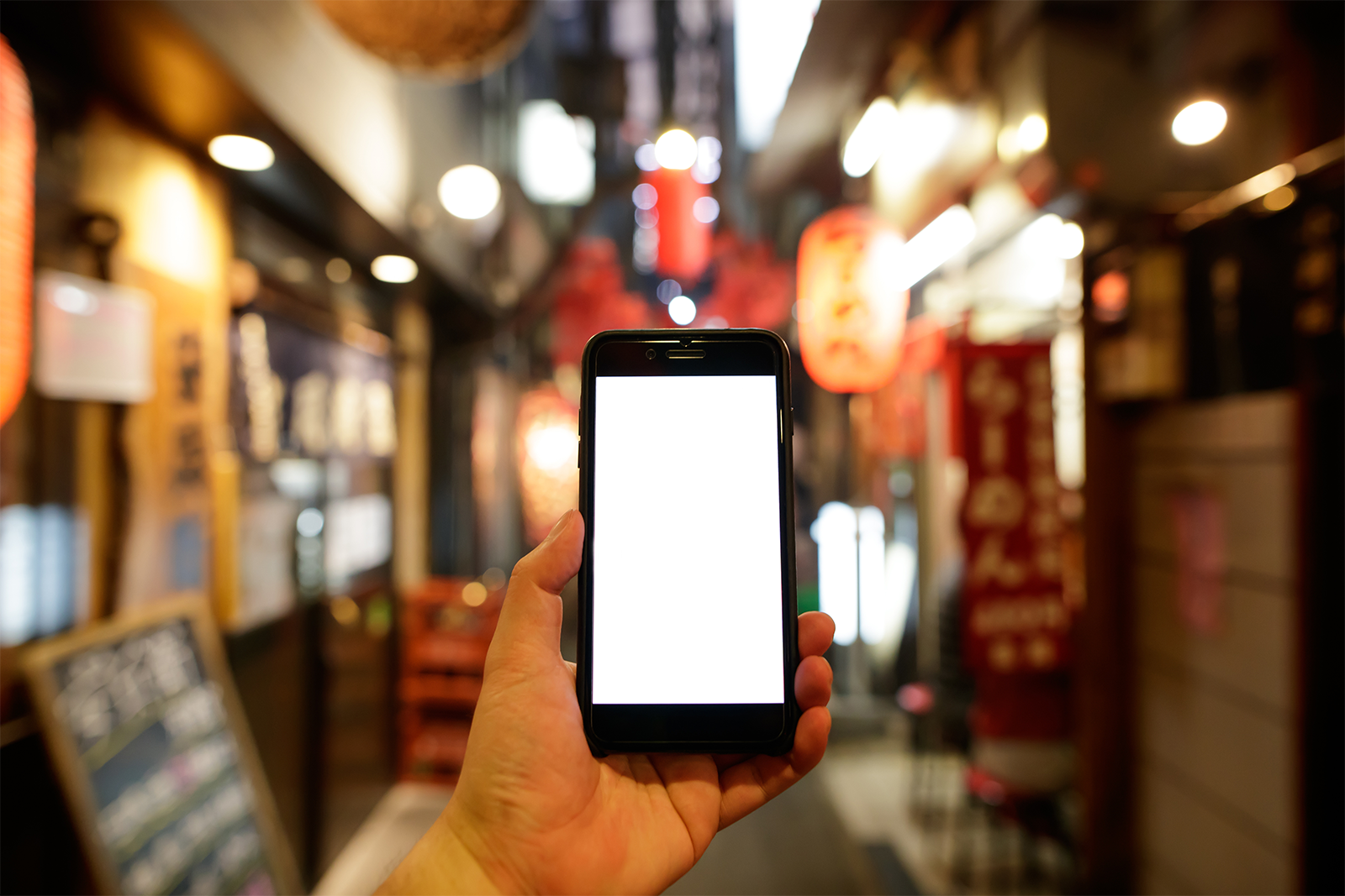 スマートフォンを持ちながら夜の商店街を歩く人の手のアップ。背景に明るい照明と看板が並び、暖かい雰囲気のアーケード街。動画によって道案内をする。