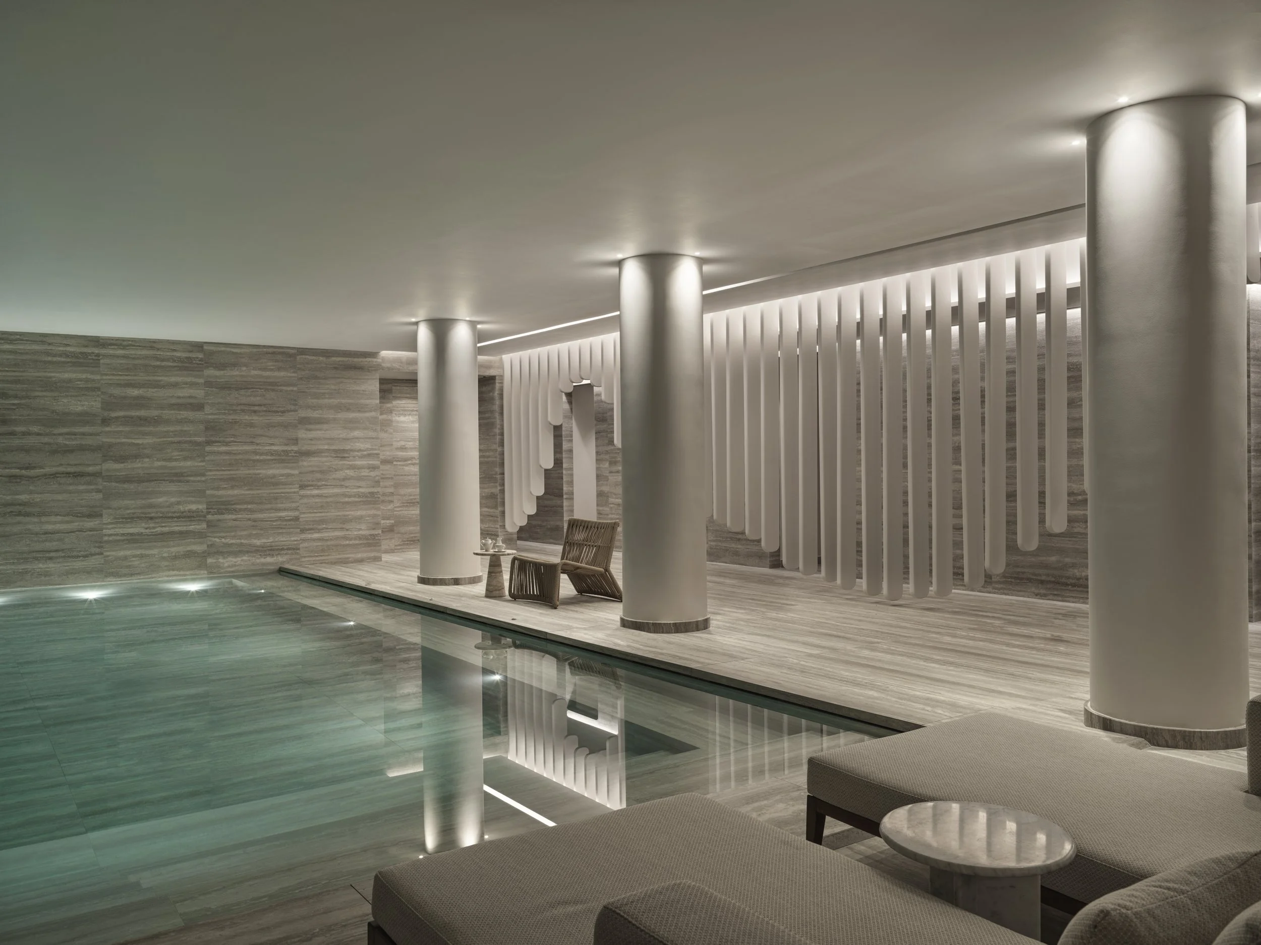 Luxury indoor swimming pool area with modern design, featuring white pillars, textured grey walls, wooden lounge chairs, and soft lighting.