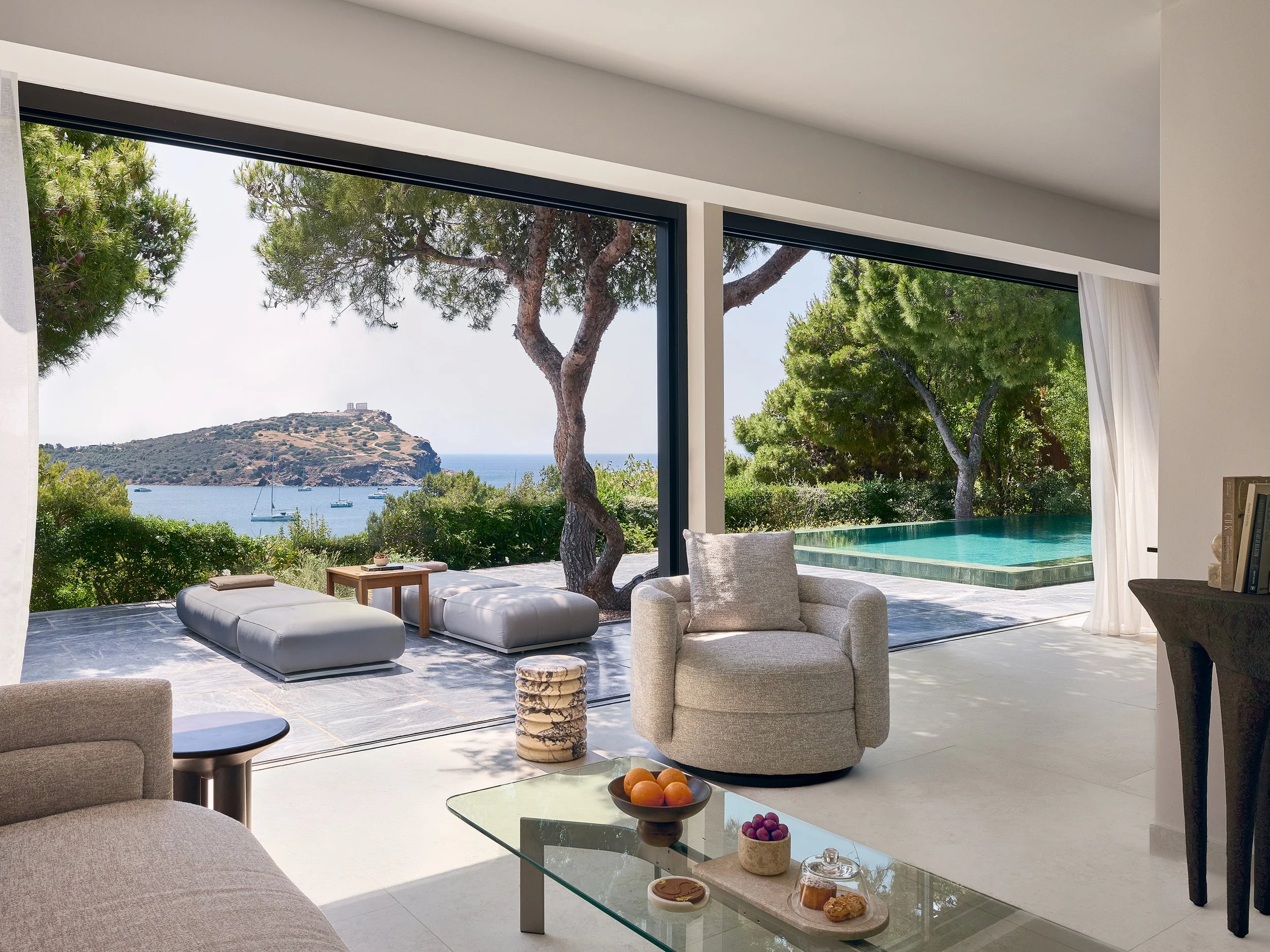 Modern living room with large glass sliding doors showing outdoor view of trees, the ocean, sailboats, and a rocky hilltop with a small building. Inside, there is a gray armchair, a glass coffee table with fruit, a beige ottoman, and a pool visible through door.