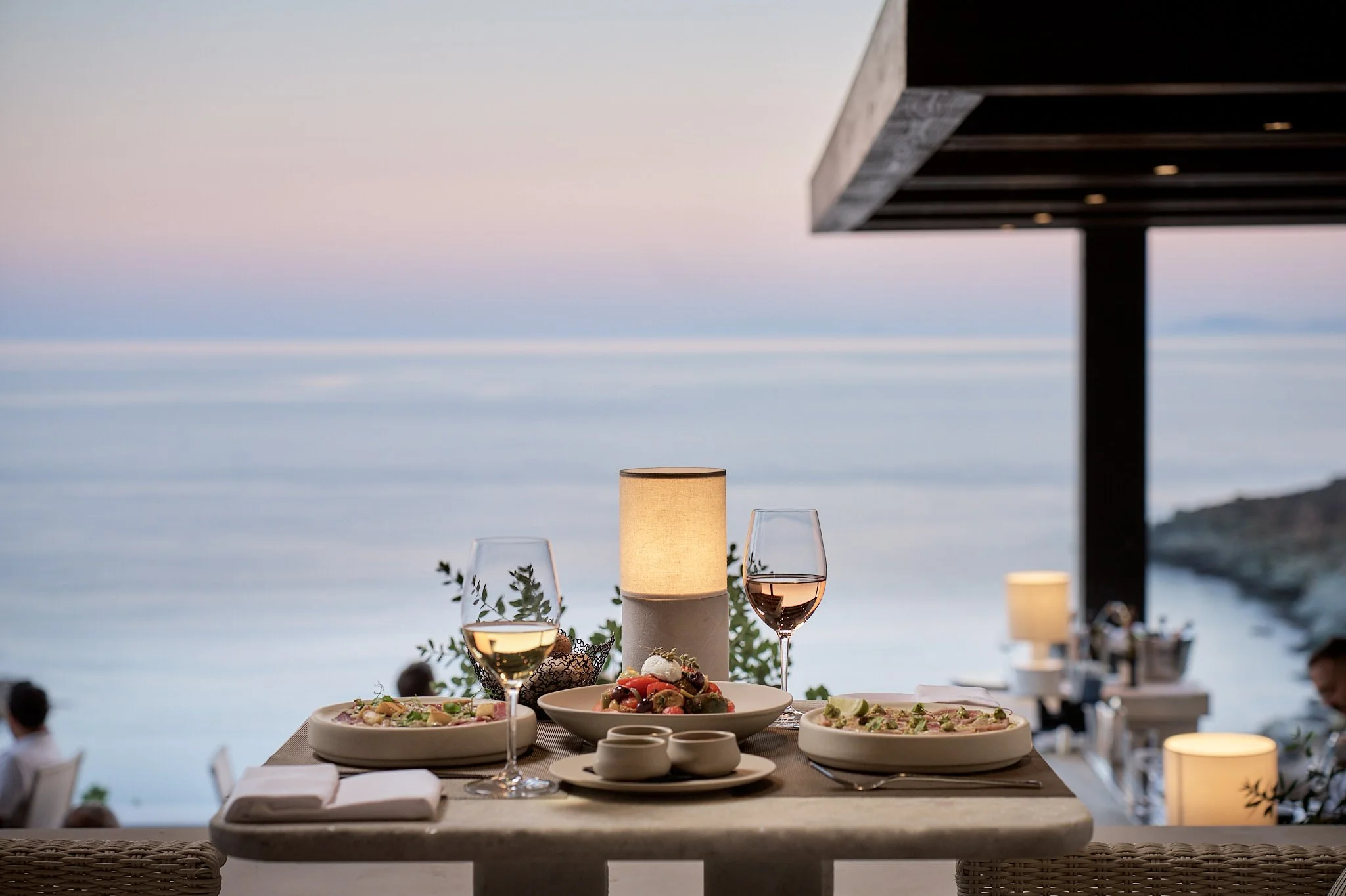 Elegant outdoor dining table set for dinner with wine glasses, a lit lamp, and a view of the ocean at dusk.