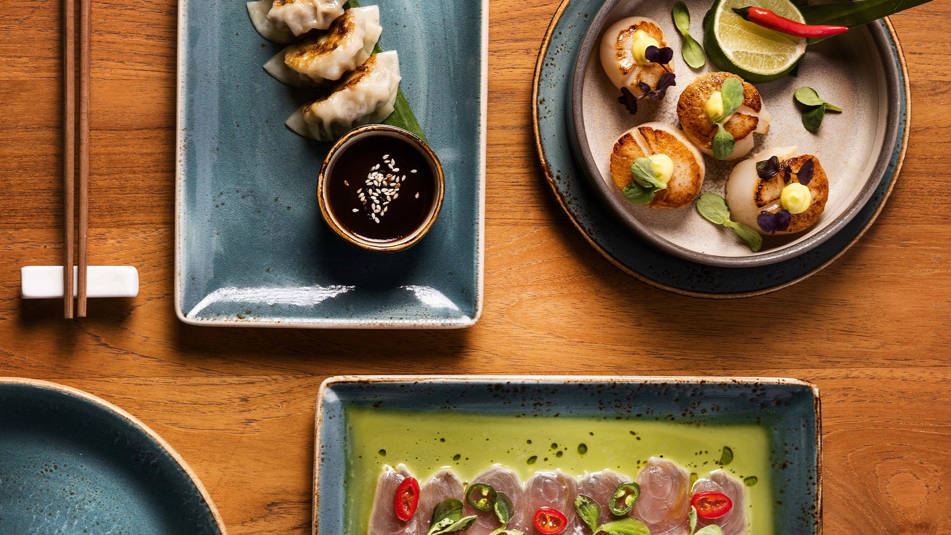 Assorted Japanese dishes on blue ceramic plates, including gyoza with dipping sauce, scallops with garnish and lime, and thinly sliced raw fish with chili peppers and green onions, on a wooden table.