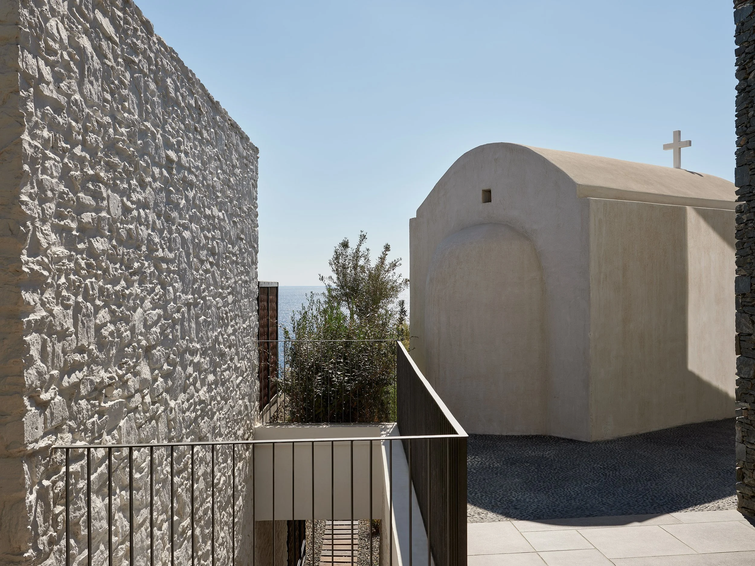 View of a small white church with a cross on top, flanked by stone walls, overlooking a body of water and some greenery under a clear blue sky.