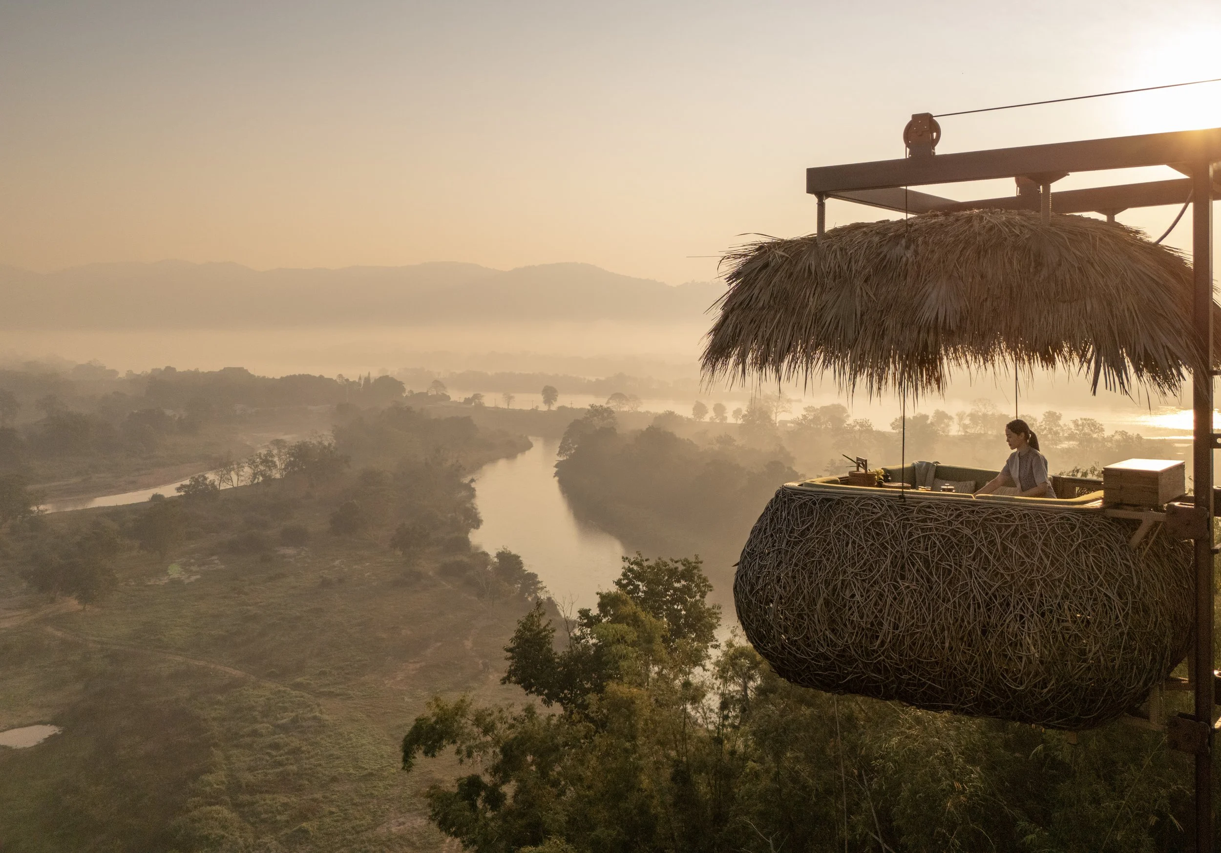 A person standing in a thatched-roof balcony overlooking a misty river and landscape during sunrise or sunset.