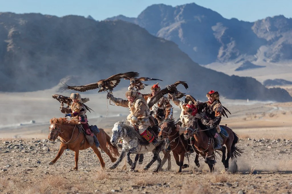 Group of people riding horses across a desert landscape with mountains in the background. Some riders are holding birds of prey.