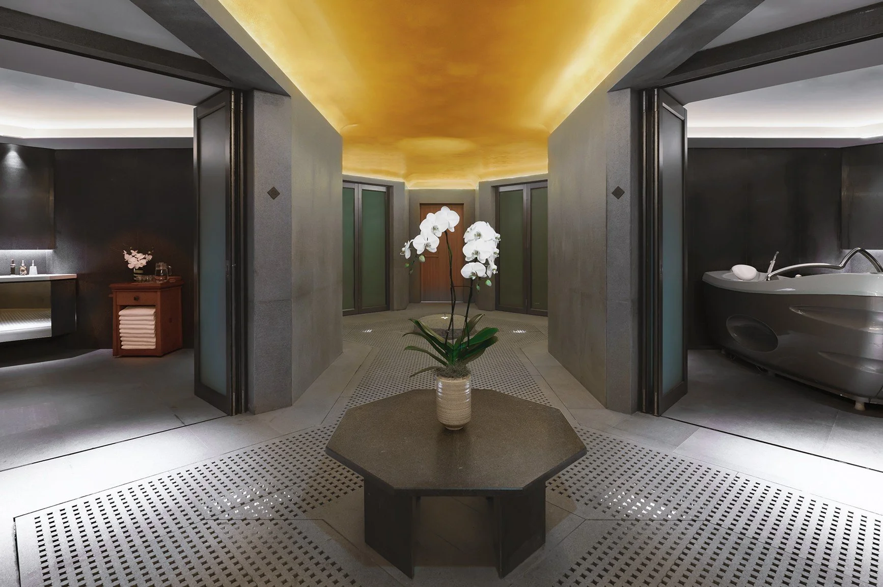 Modern spa lobby with central orchid arrangement, gold ceiling, and separate areas for a hot tub and massage table.