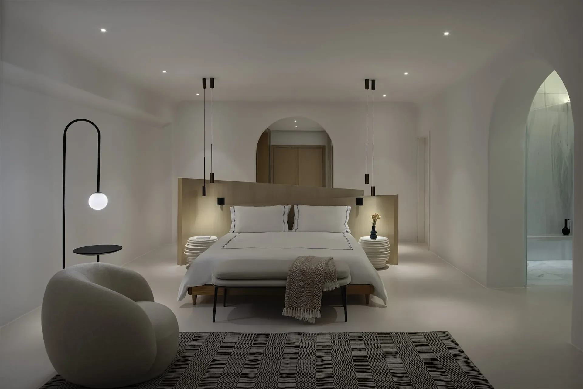 A minimalist bedroom with white walls, a white bed with black trim, and a beige headboard. There are two white bedside tables with black vases and yellow flowers. A unique black floor lamp with a globe-shaped light is on the left, and an arched doorw