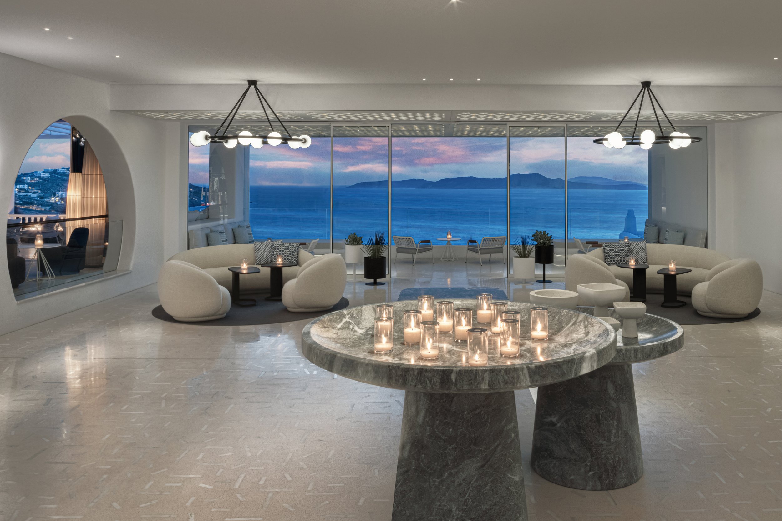 Elegant modern living room with large floor-to-ceiling windows offering a view of the ocean and distant islands, featuring white curved sofas, black side tables with candles, a round marble table with lit candles, and minimalist decor.