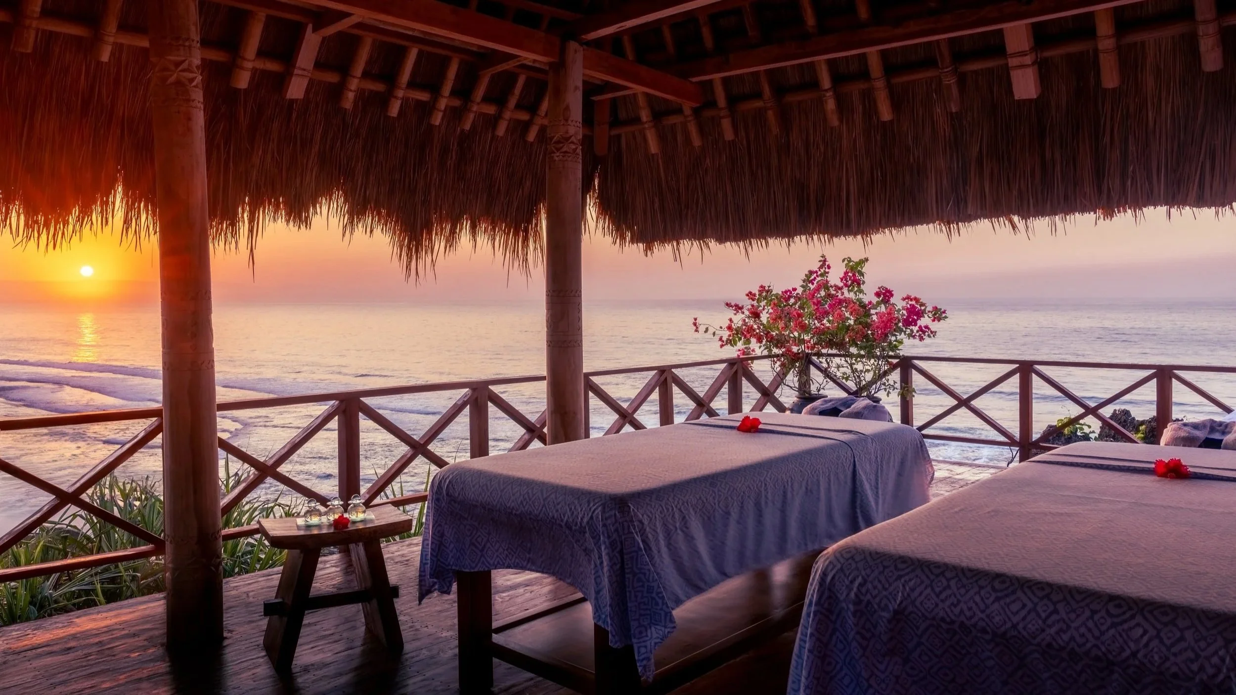 Beachside wellness or massage cabana with two massage tables, colorful flowers, and a sunset over the ocean.
