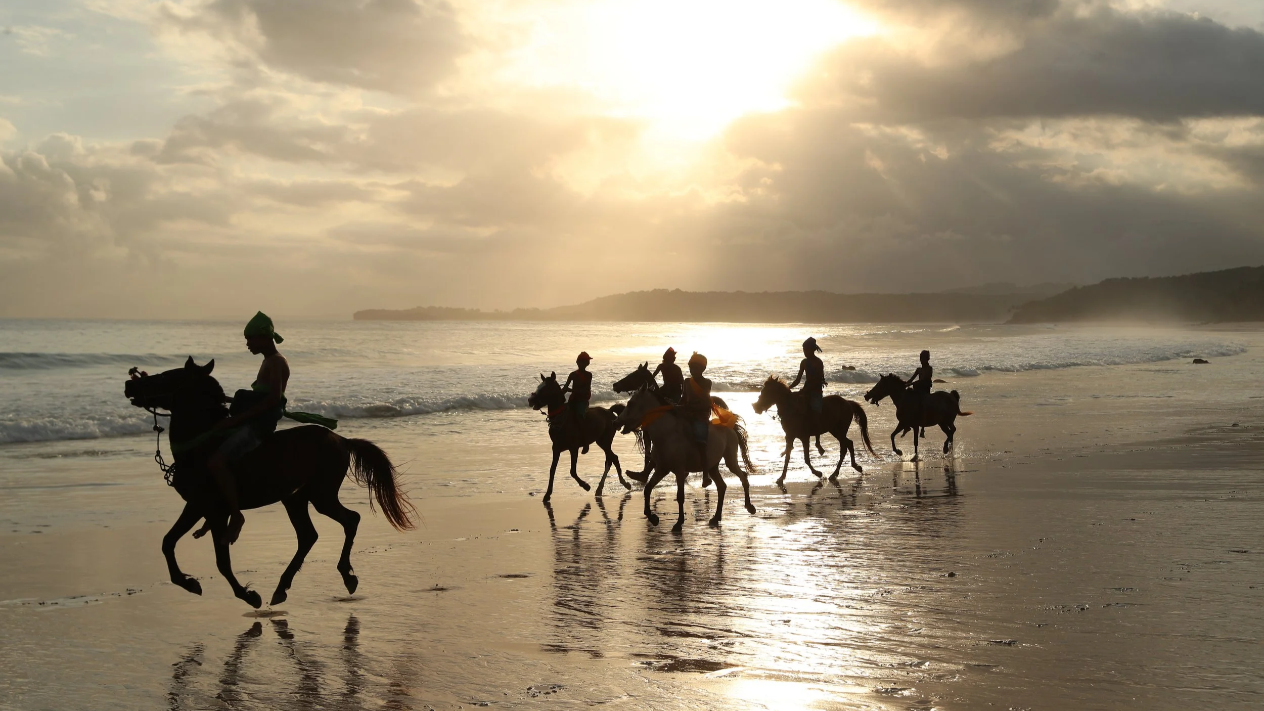Silhouettes of people riding horses along the beach during sunset, with waves crashing and hills in the distance.