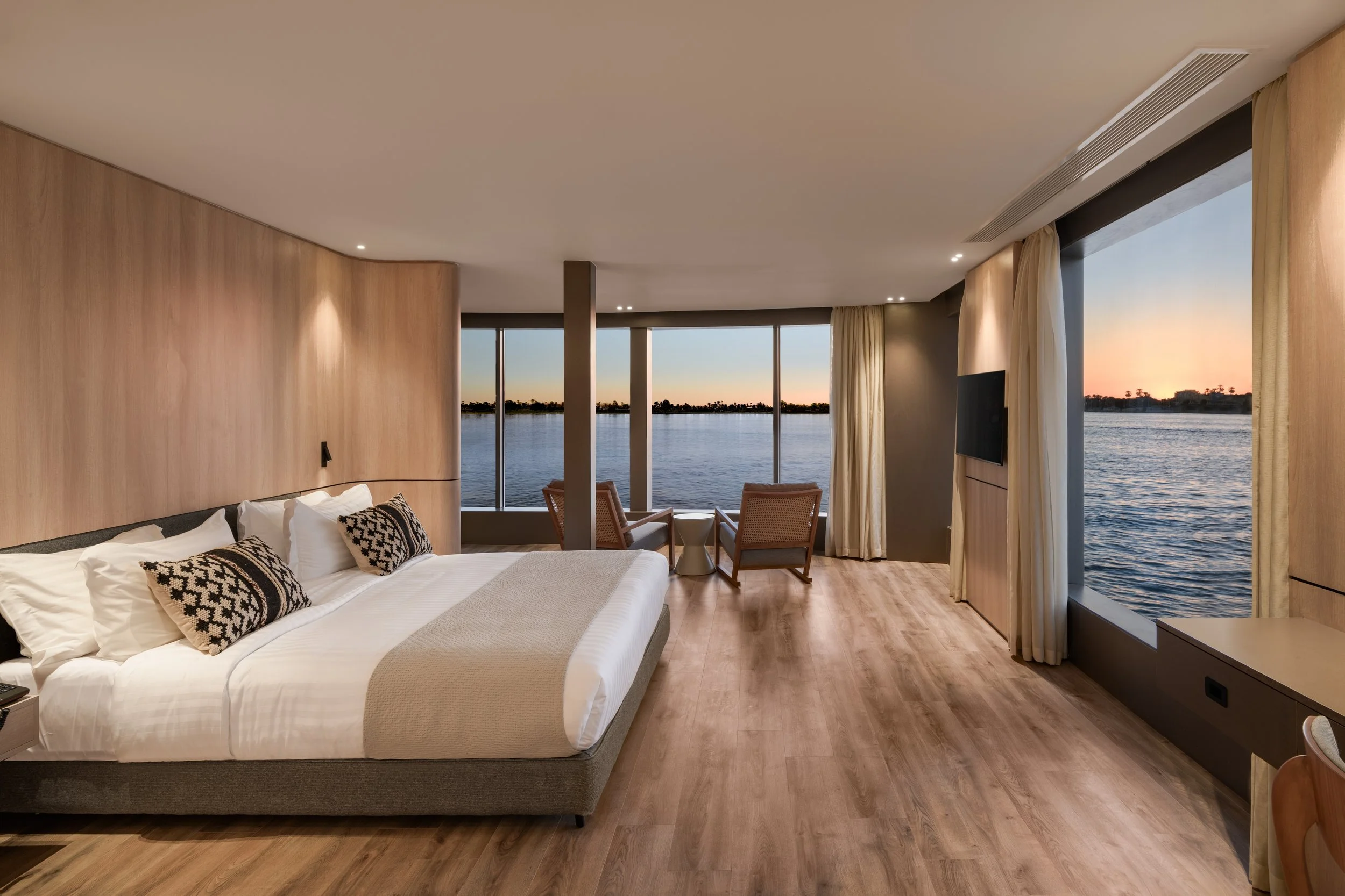 Modern room with a view of a river at sunset, containing a bed with white linens, decorative pillows, a sitting area with two chairs and a small table, large windows with curtains, and wooden flooring.