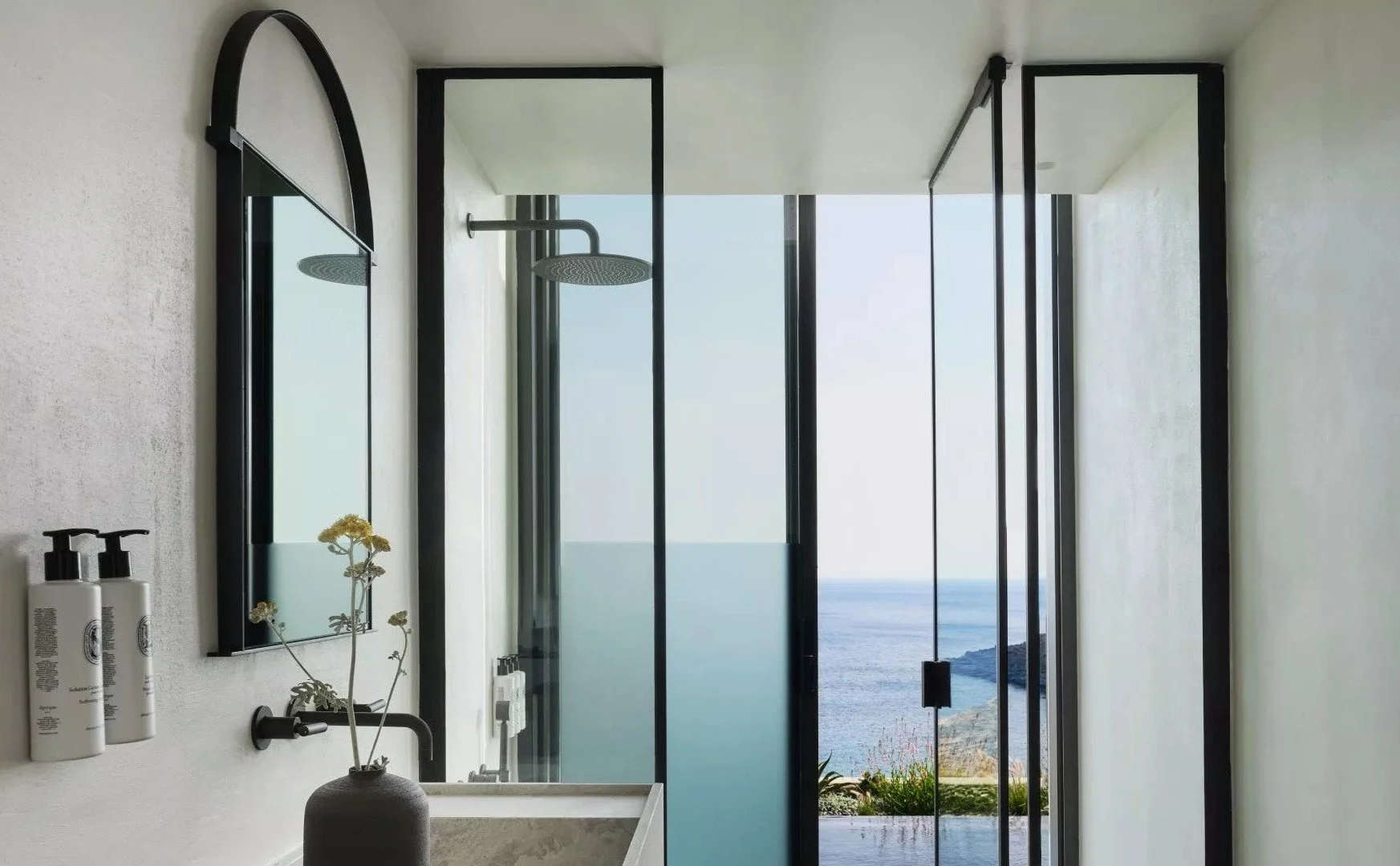 Modern bathroom with a shower area overlooking the ocean through floor-to-ceiling glass doors, minimalistic decor, a mirror, and toiletries on the wall.