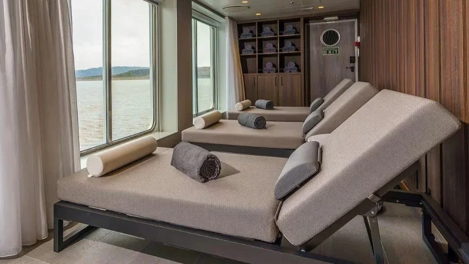 Two lounge chairs with rolled towels and cushions, positioned near large windows overlooking a body of water, with wooden wall paneling and a built-in cabinet in the background.
