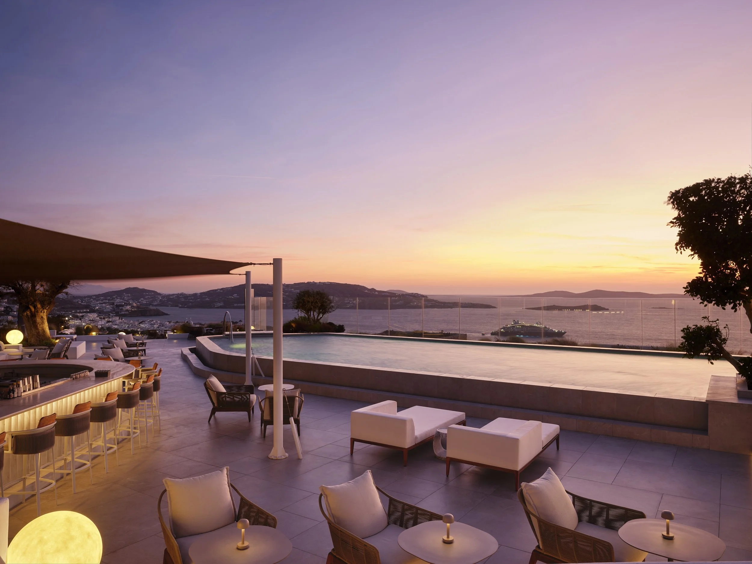 Luxury outdoor pool area on a terrace overlooking the ocean at sunset, with lounge chairs, a bar, and umbrellas.