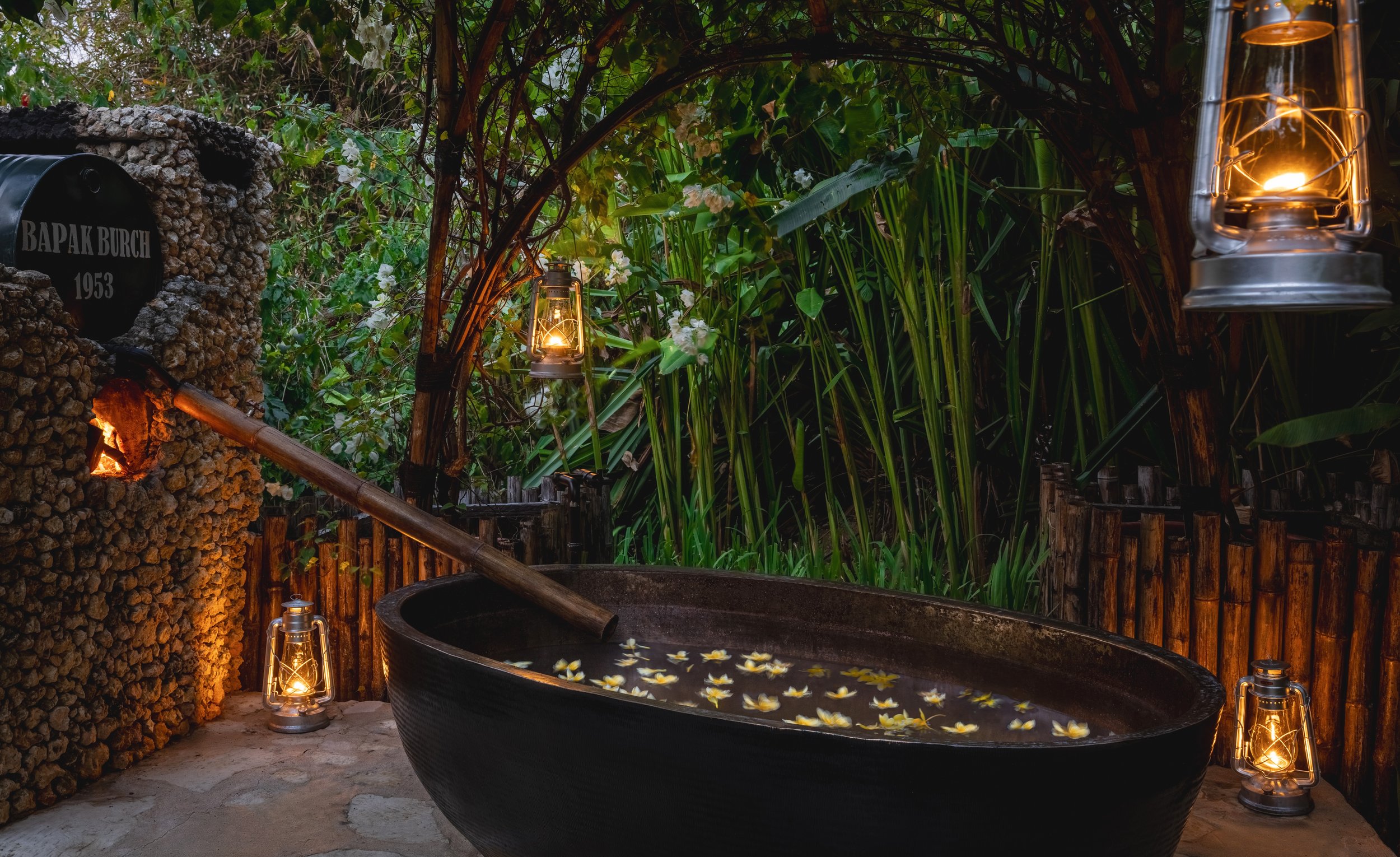 Outdoor tub filled with water and floating white flowers, surrounded by lanterns and lush greenery with bamboo fencing.