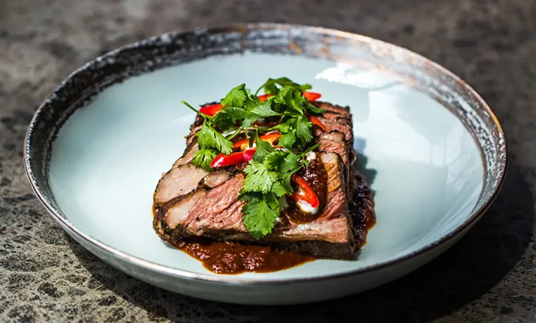 A plate of sliced grilled steak topped with chopped red chili peppers and cilantro garnishes, served with sauce on a light-colored ceramic plate.