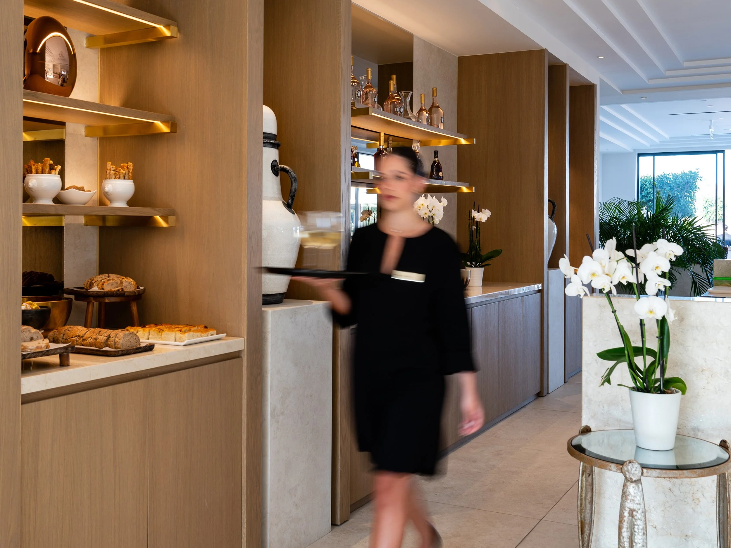 A well-lit hotel lobby or restaurant with wooden shelves displaying decorative items and baked goods, orchid plants, and large windows in the background.